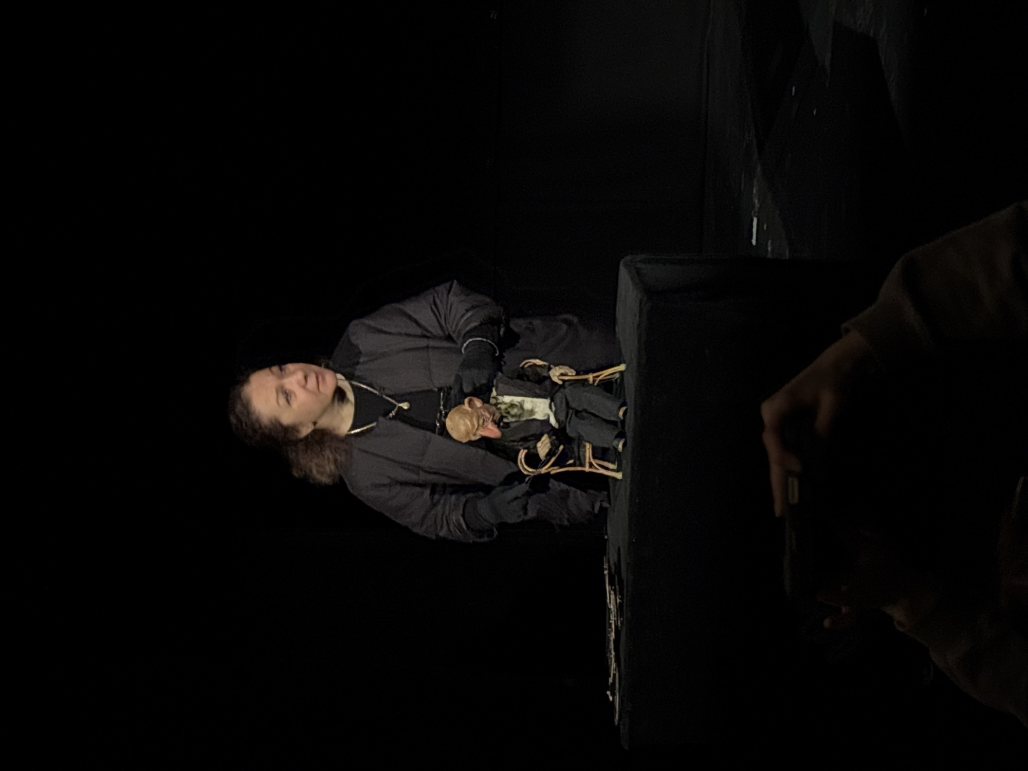 The image shows a person in a dark environment, possibly a theater or performance space. The person is wearing a dark jacket and gloves, and they are holding a puppet or doll. The puppet appears to have a human-like face. Another person is partially visible in the lower right corner, holding a phone as if taking a picture. There are dark, undefined objects in the background. The location and details are insufficient to determine if the scene is in Tbilisi, Georgia.