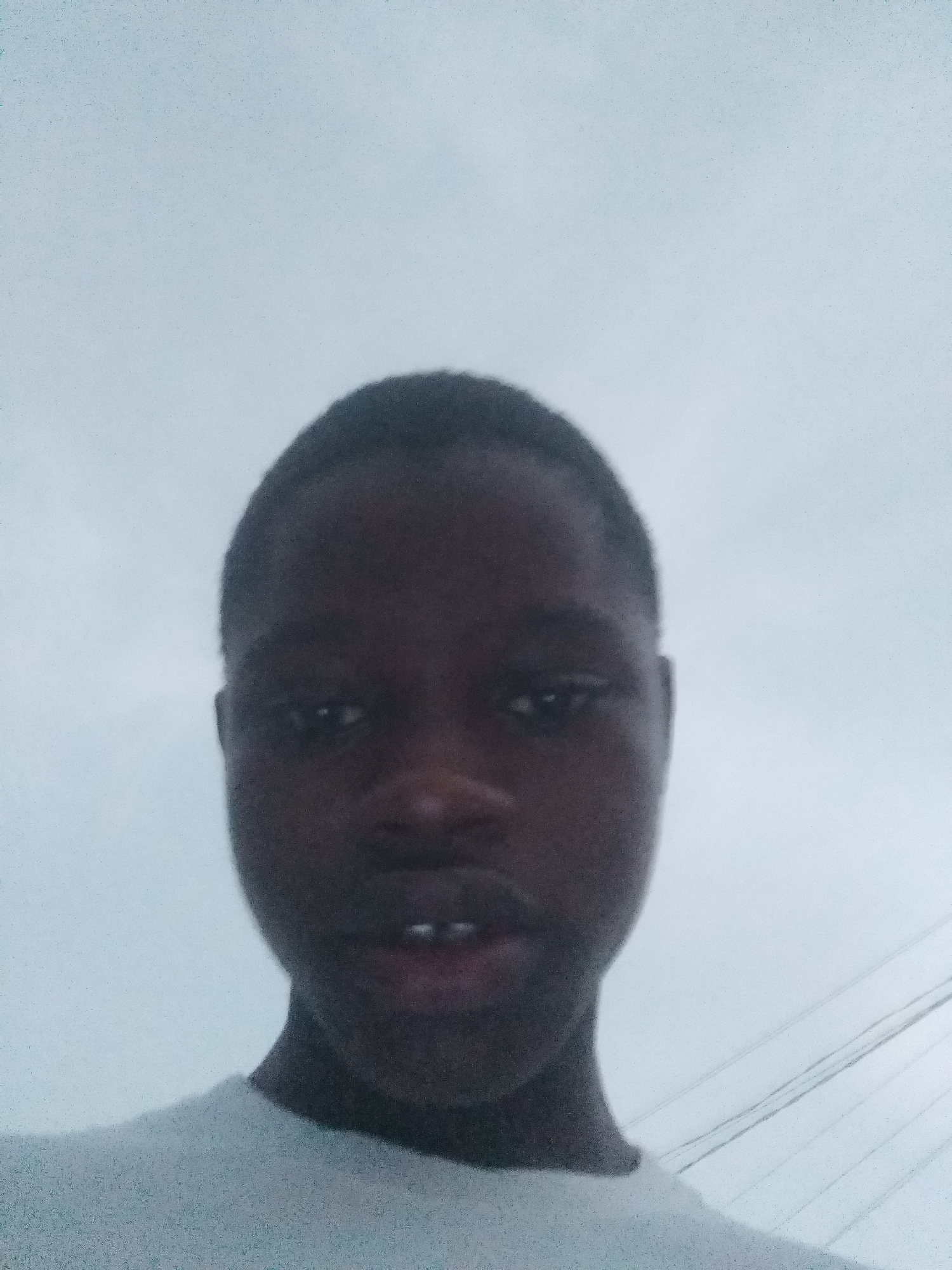 The media file features a close-up portrait of a young person, likely male, with dark skin and short, dark hair. The individual's face is centered and prominent in the frame, looking directly at the viewer with a neutral to slightly serious expression. Their mouth is slightly open, revealing a gap between the two front teeth. They are wearing a light-colored (possibly white or light grey) t-shirt or top, visible at the bottom of the frame around the neck and shoulders.
The background is a uniform, bright, light grey sky, indicating an overcast or cloudy daytime. In the lower right corner, several parallel utility or power lines are subtly visible against the sky. The setting appears to be outdoors in Shangisha, Nigeria, possibly in an urban or suburban area. No specific activity is depicted other than posing for the camera. There is no visible text in the image.