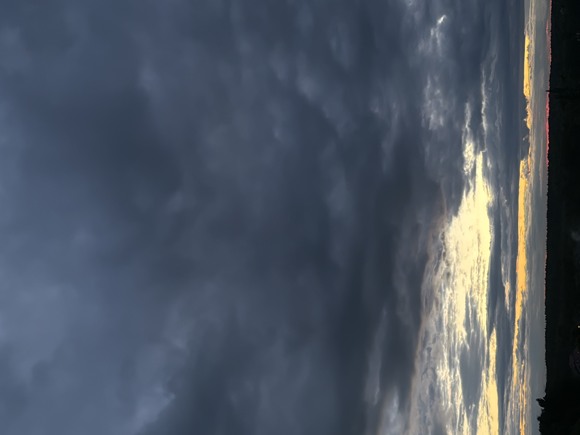 The image depicts a sky dominated by dark, stormy clouds. A sliver of sunset is visible along the horizon, with shades of orange and yellow peeking through the cloud cover. The horizon appears to be lined with trees. The location and city are unknown.