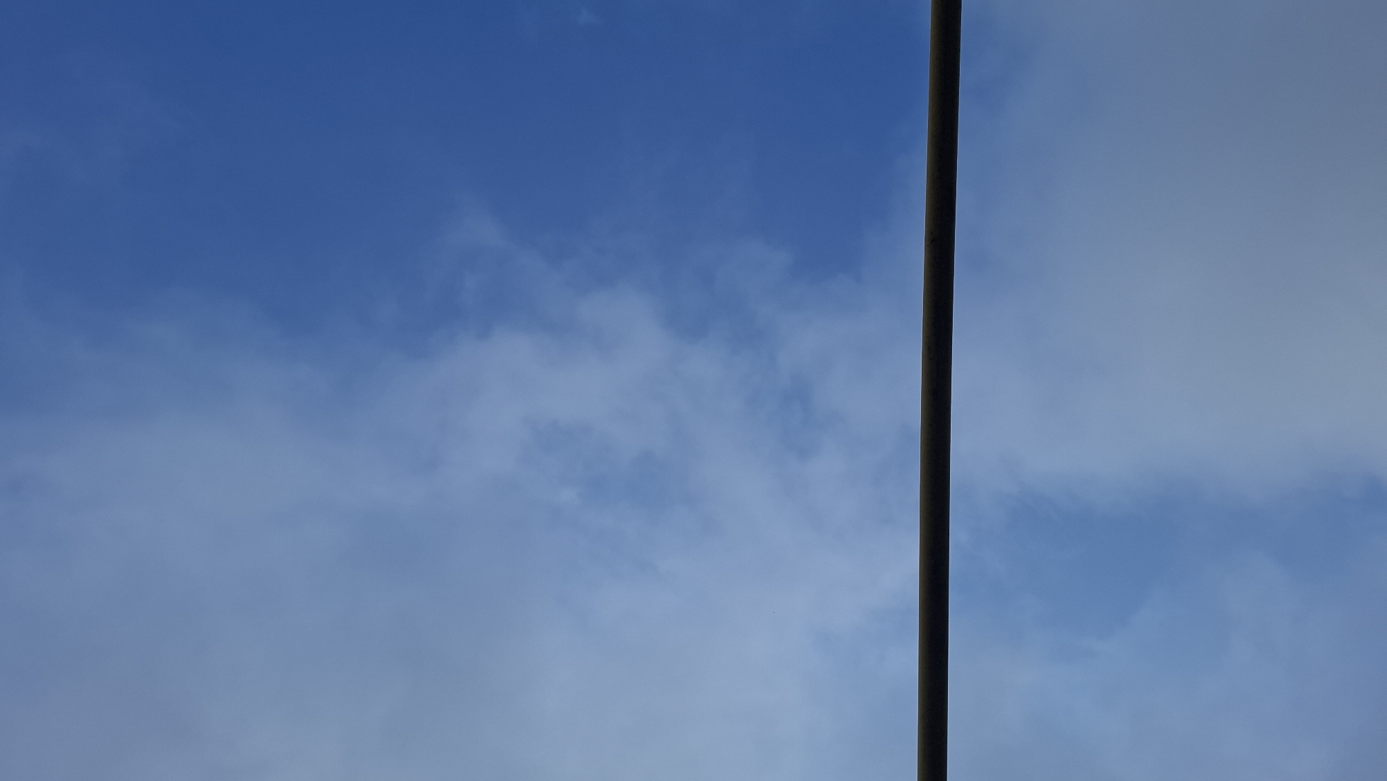 The image shows a mostly clear blue sky with scattered white clouds. A vertical gray pole bisects the right side of the frame. The location is uncertain, and there are no people, specific subjects, objects beyond the pole and clouds, actions, or interactions depicted in the image.