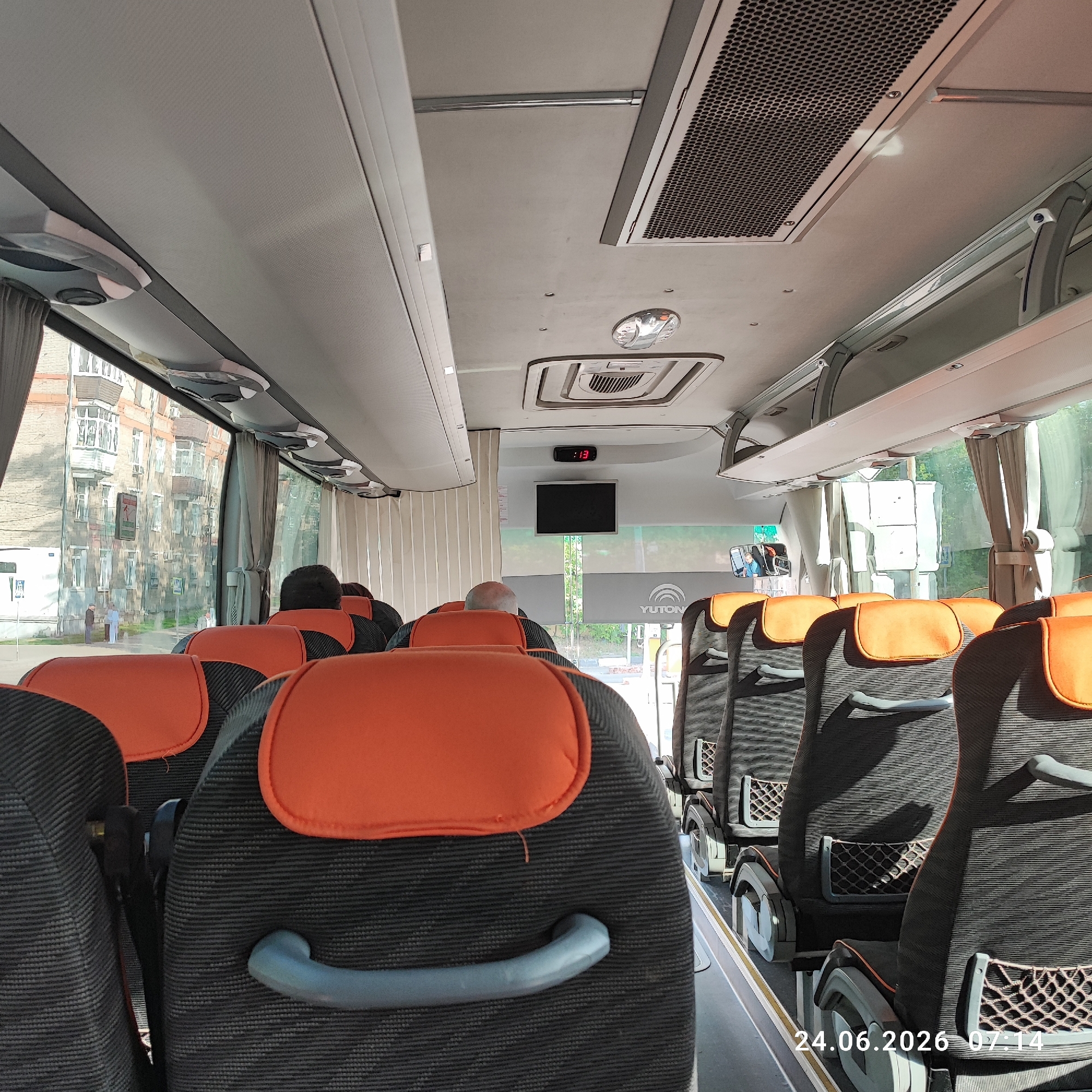 The image shows the interior of a bus, looking towards the front. The seats are upholstered in dark fabric with orange headrests and have gray handles. Some seats are occupied by passengers, their heads and shoulders visible. The bus interior features overhead compartments, air vents, and lighting fixtures. A small screen and a digital clock displaying "13:" are mounted on the ceiling. Through the windows, buildings and trees are visible, suggesting an urban or suburban setting. The timestamp "24.06.2026 07:14" is superimposed on the lower right corner of the image, indicating the time and date. The scene appears to be a passenger bus in motion during the morning. The logo "YUTON" is visible on a banner inside the bus near the front.