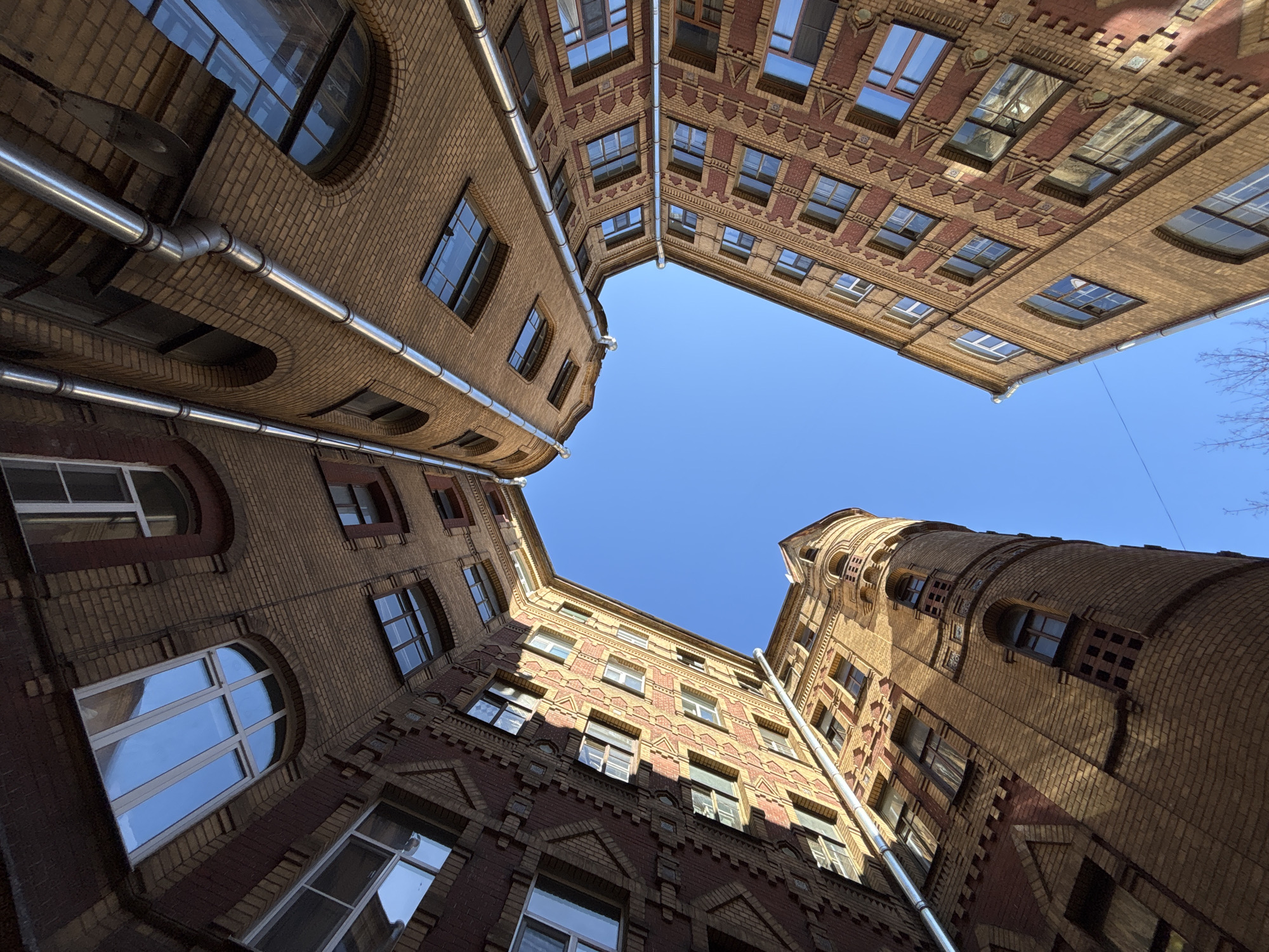 The image captures a courtyard in Saint Petersburg, Russia, viewed from a low angle looking upwards. Four buildings enclose the courtyard. The buildings are constructed of reddish-brown and light yellow brick, featuring arched windows and decorative brickwork. Metal pipes run along the exterior walls. The sky above is clear and blue.
