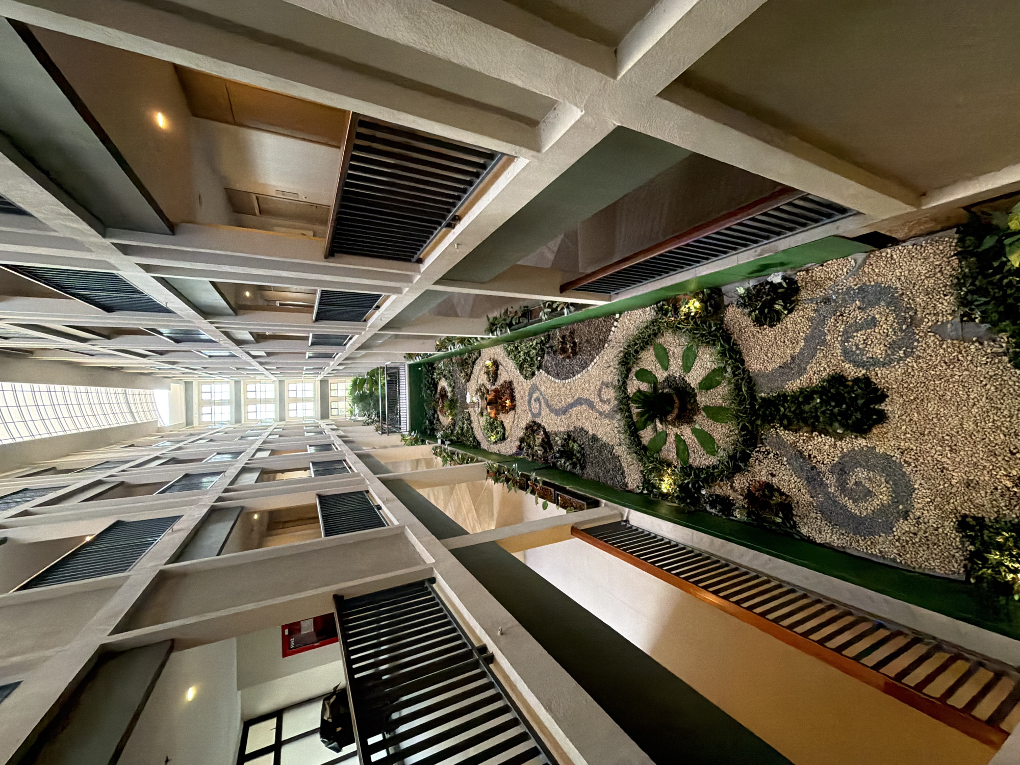 The image captures an interior view of a multi-story building with an atrium that features a long, decorative garden bed running down the center. The garden is adorned with gravel, plants, and a small water feature. The atrium has balconies on each side, with railings and doorways leading to rooms. The building's architecture is modern, with concrete columns and beams. The ceiling is a skylight, allowing natural light to filter in. The overall impression is one of a well-maintained, indoor green space within a hotel or residential complex. The location is identified as Bang Muang, Thailand.