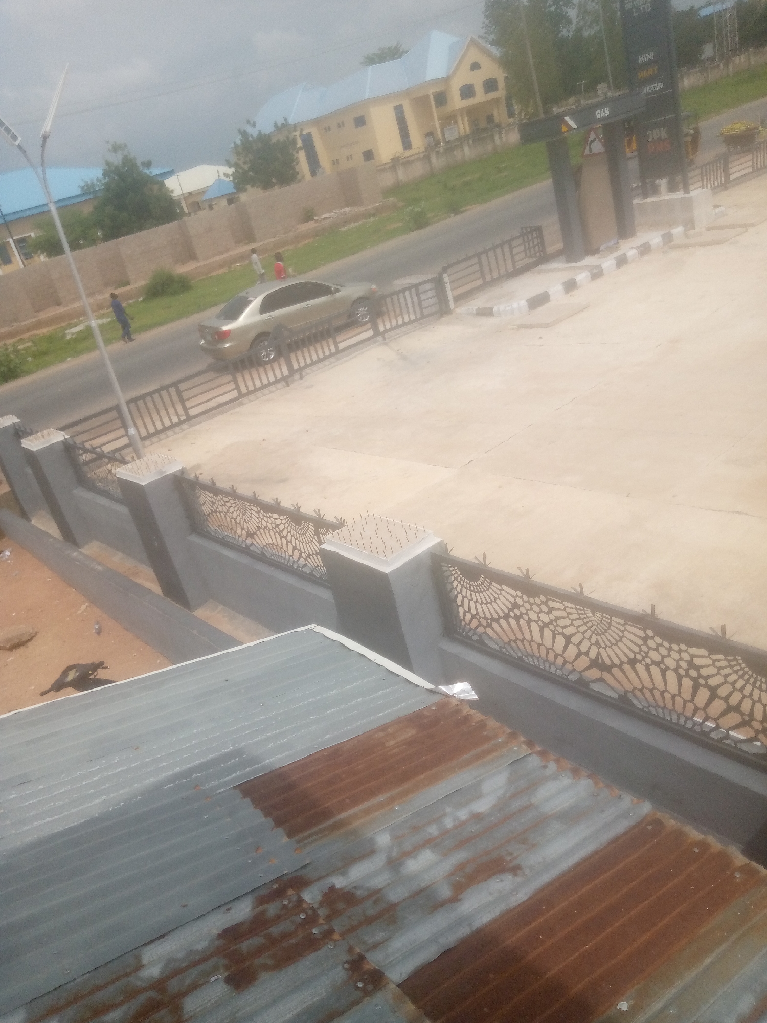 The image captures an exterior scene in Jalingo, Nigeria, focusing on a roadside area. In the foreground, a corrugated metal roof, showing signs of rust, is partially visible. Beyond the roof, a grey concrete wall with decorative metal railings topped with spikes separates the foreground from the street. A silver sedan is parked on the right side of the road, which is paved and has a sidewalk.
Further down the street, a gas station canopy with signage for "JPK PIMS" and "GAS" is present. Behind the gas station, a yellow building with a blue roof and a smaller white building with a blue roof are visible. The sky is overcast. Two individuals, appearing to be men, are walking on the sidewalk near the silver sedan. One is wearing a blue shirt and dark pants, while the other wears a red shirt and dark shorts. Another individual, dressed in dark clothing, is walking further down the road. A street light with solar panels is situated on the left side of the image. The overall environment suggests an urban or semi-urban setting.