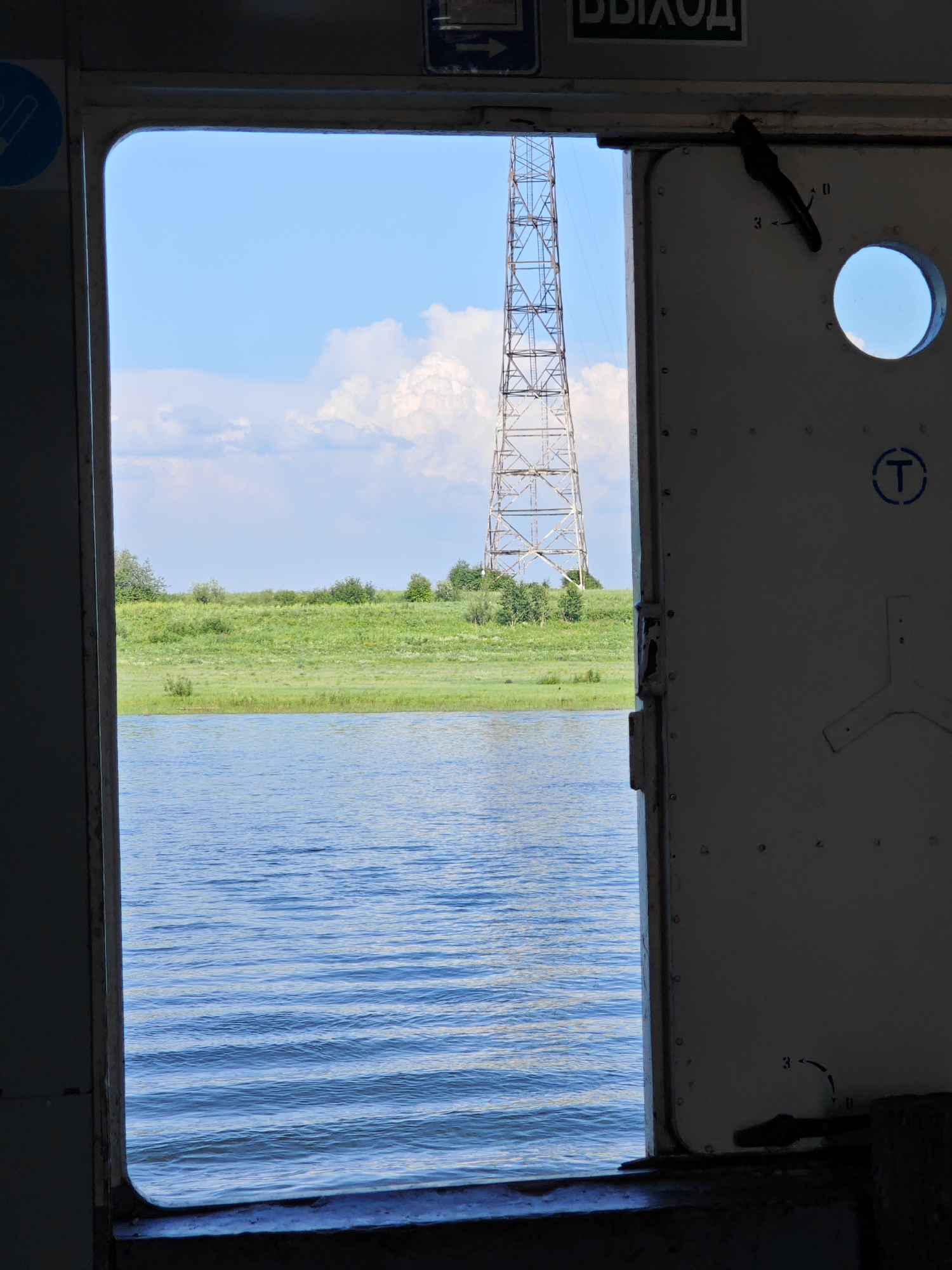 The image shows an open doorway of a vessel, looking out onto a body of water and a grassy embankment. A tall, lattice-like tower, possibly a radio or power pylon, is visible in the distance on the embankment, beyond a line of trees. The sky is blue with scattered white clouds. The doorway itself is framed by dark metal, and the interior of the vessel, visible on the right, appears to be metallic with some visible rivets and a circular viewing port. A latch mechanism is visible on the door. The scene appears to be in Verkhnepashino, Russia, given the context. There are no people visible in the image.