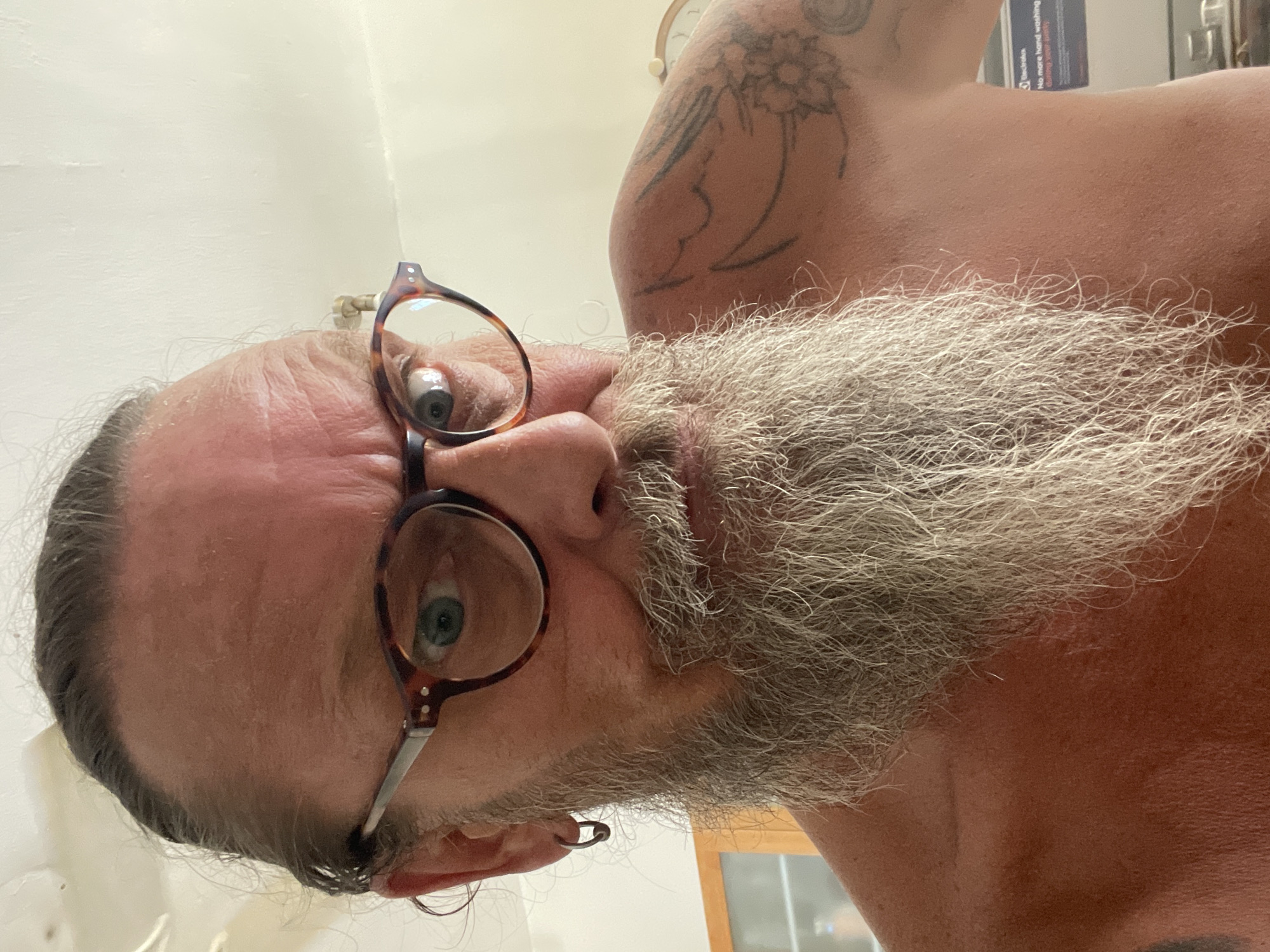 A man with graying hair pulled back, a long gray beard, and blue eyes is looking directly at the camera. He is wearing tortoise-shell glasses and a hoop earring in his left ear. He has a tattoo on his right shoulder. The man is shirtless and appears to be indoors in a room with white walls and a ceiling. A blurred background shows a cabinet and possibly a washing machine. The lighting suggests it is daytime. The image was taken in Netanya, Israel.