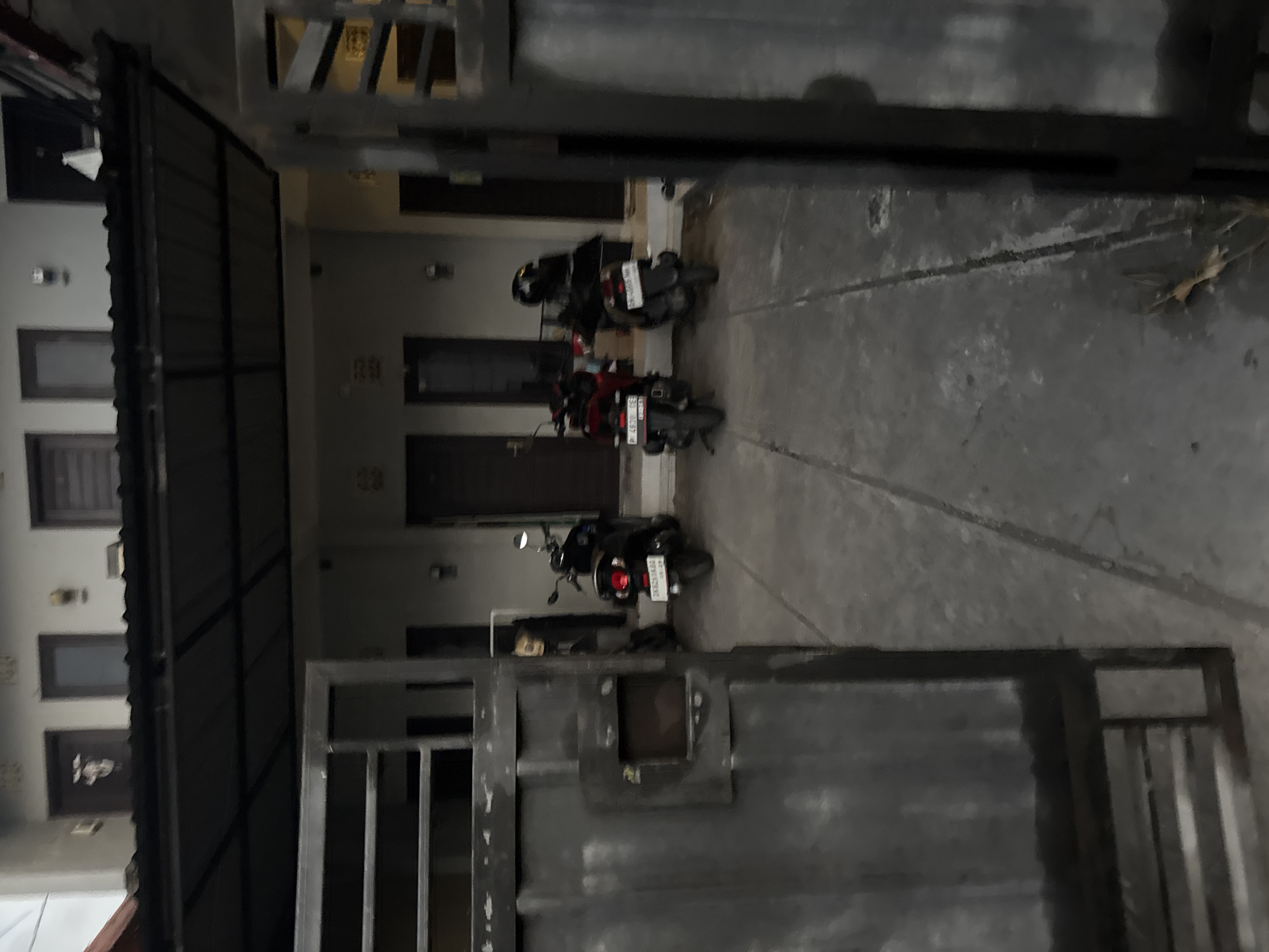 The media file shows an outdoor scene in Sukawati, Indonesia, likely in a residential or commercial complex with multiple entrances. In the foreground, a large, dark metal gate is partially visible, with vertical slats and a square opening. Behind the gate, the scene opens up to a pathway or driveway with a concrete surface.
Several motorcycles are parked along the pathway. One is a dark-colored scooter with a helmet on its seat. Further down, a red motorcycle is parked, also with a helmet. The motorcycles suggest that this is a place where people regularly use them for transportation. The architecture of the building visible includes dark-framed windows and doorways. The overall lighting suggests it might be late afternoon or early evening, as there are shadows but the scene is not fully dark. The setting appears to be a covered area or a walkway adjacent to buildings. There are no people visible in the image. One of the motorcycle license plates has text that is partially legible: "P 4650 GSN". Another license plate appears to be "MR 9810 KB".