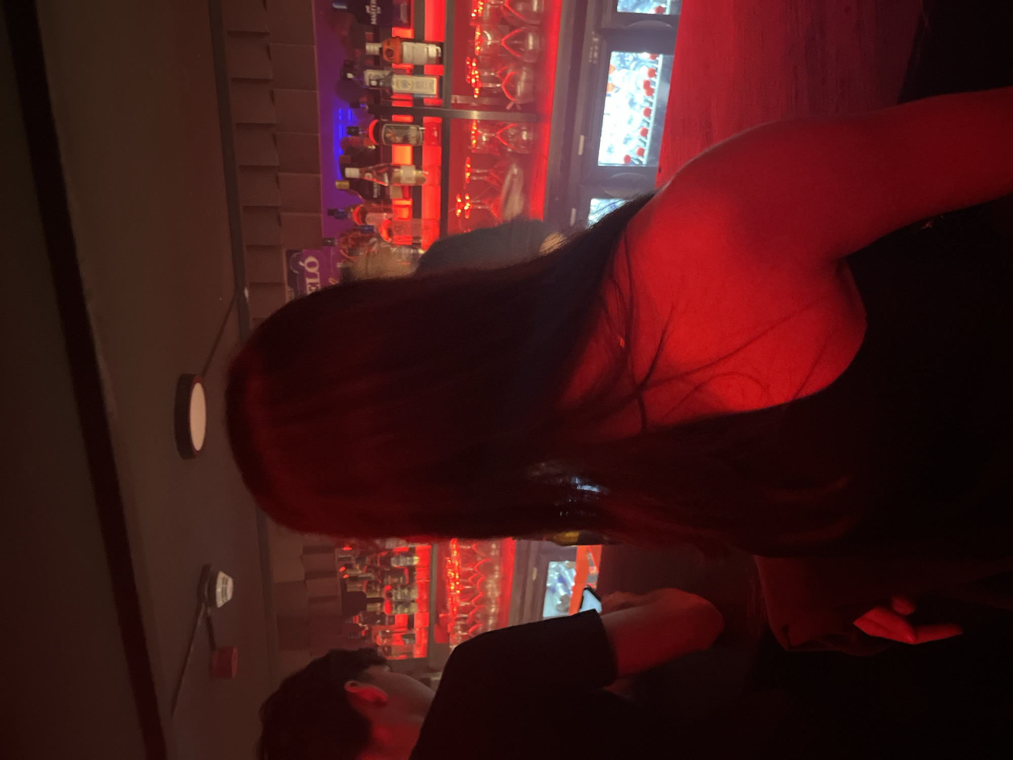 A person is standing in a bar or club in Valencia, Spain. Behind them is a bar stocked with bottles and glasses, illuminated with red light. The location is likely in a dark area with light sources of varying colors. Another person is partially visible, facing the right side of the frame. The light casts shadows and obscures the details of the people and their actions.