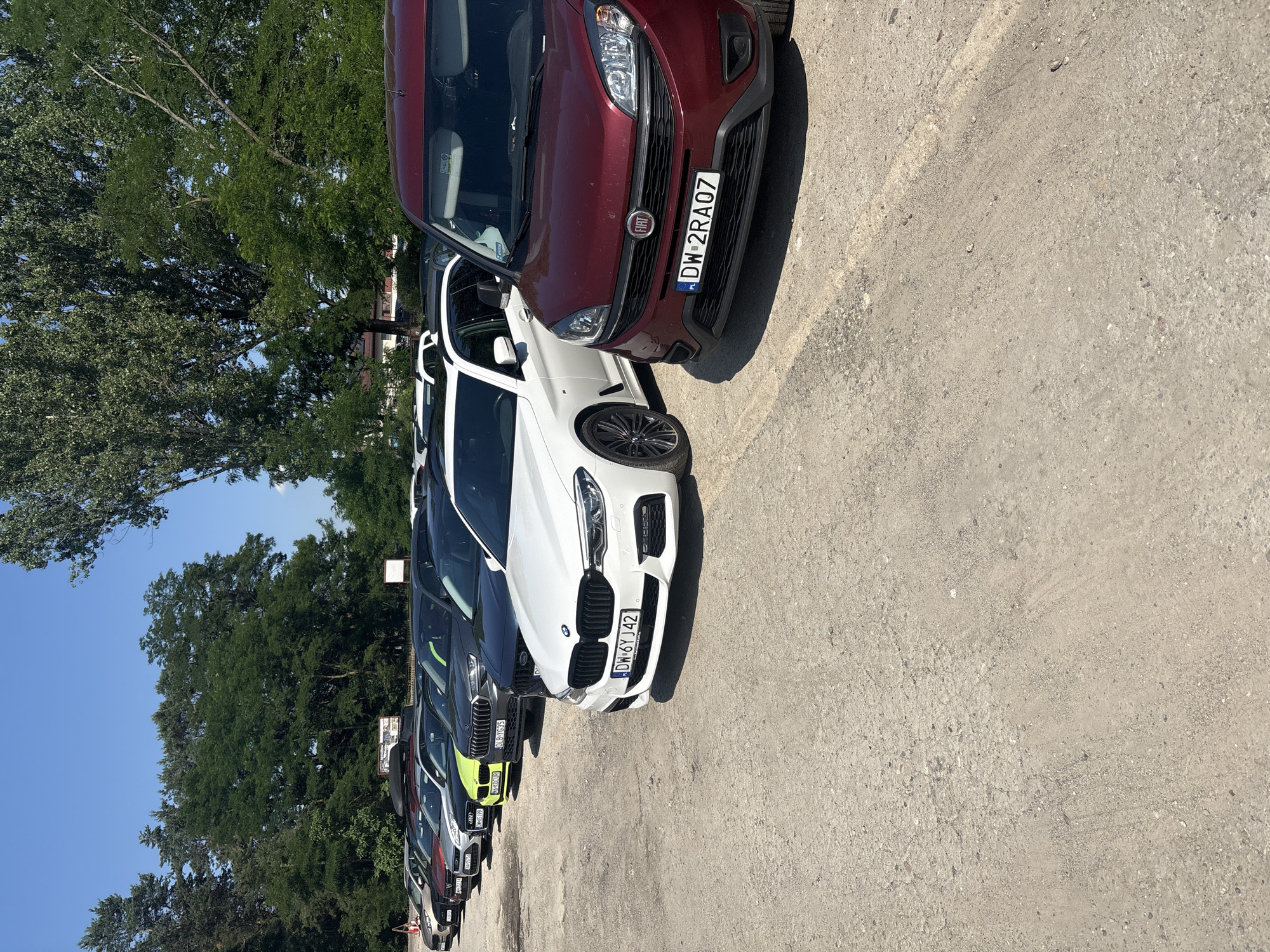 In the city of Kobyla Góra, Poland, on a clear, sunny day, a line of cars is parked on a gravel surface. The foreground features the front of a dark red Fiat van, displaying a license plate that reads "DW 2RA07". To its left, a white BMW station wagon is parked, its front bumper and license plate visible. The license plate on the BMW shows "DW 6Y J42". Behind the BMW, a row of other vehicles is visible, including a dark gray car with a lime green accent and other dark-colored cars. In the background, a line of green trees stands tall against a bright blue sky. The overall scene depicts a parking area in a semi-rural or suburban setting.