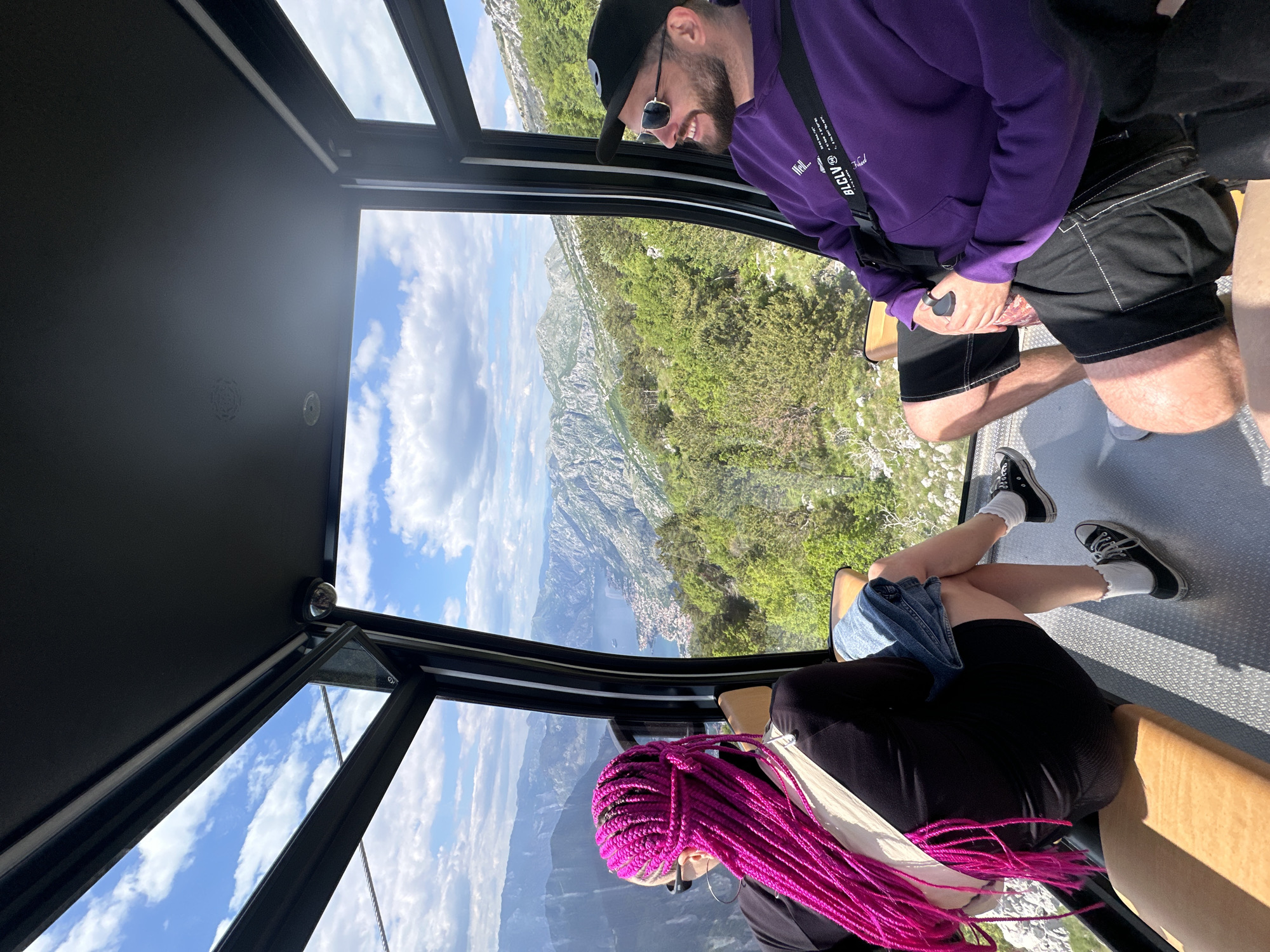 The image shows an interior view of a gondola car, likely ascending or descending a steep incline, with a scenic view of Škaljari, Montenegro, visible through the windows. Three people are present inside the car. A man in a purple hoodie and a black baseball cap is standing near the window, looking out and smiling. His attire includes black shorts and a strap across his body. The lower half of another man is visible, he is wearing black shorts, white socks, and black shoes. The last person is sitting and can be identified by their long, bright pink braided hair. The view from the gondola features a landscape of green mountains, a body of water (likely part of the Bay of Kotor), and possibly a distant cityscape. The gondola car itself appears modern with black frames and clear windows. The lighting is bright, suggesting it is daytime.