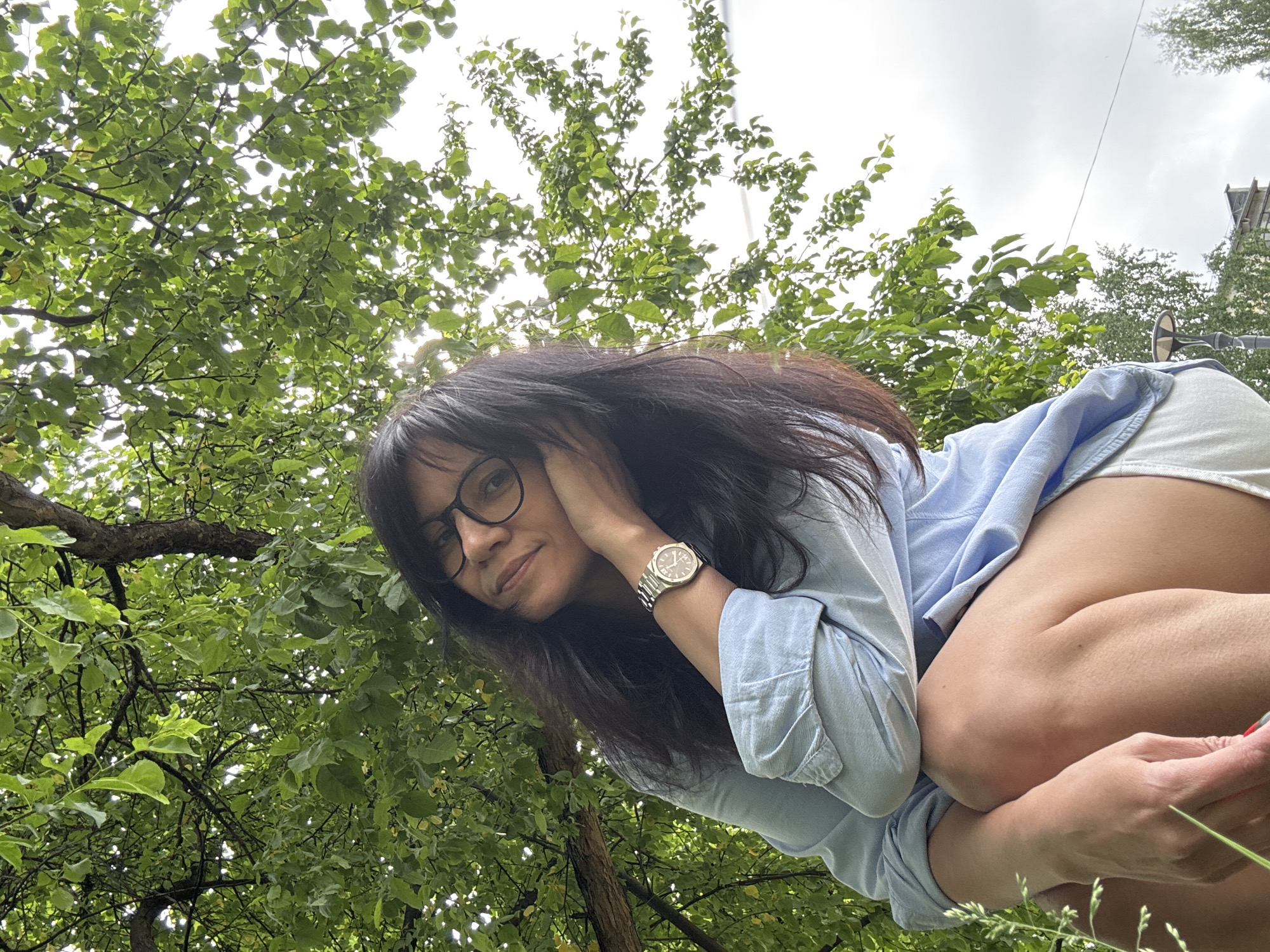 A low-angle medium shot features a woman with long dark hair and glasses, wearing a light blue button-down shirt, possibly in a park or natural area. She is looking slightly down at the camera, with her right hand near her face, possibly touching her hair, and wearing a silver wristwatch on her left wrist. The background is filled with green foliage, suggesting trees overhead, and a glimpse of the sky is visible through the leaves. The location is suggested by the surroundings and could be a park in the city of Unknown, Unknown.