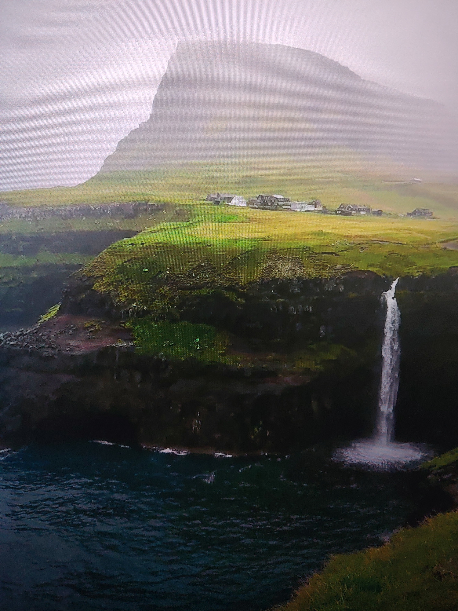 The image depicts a coastal landscape with a waterfall cascading into a body of water, likely the ocean. A small settlement or village is visible on the grassy terrain above the cliffs. A large, misty mountain looms in the background. The location is unidentified.