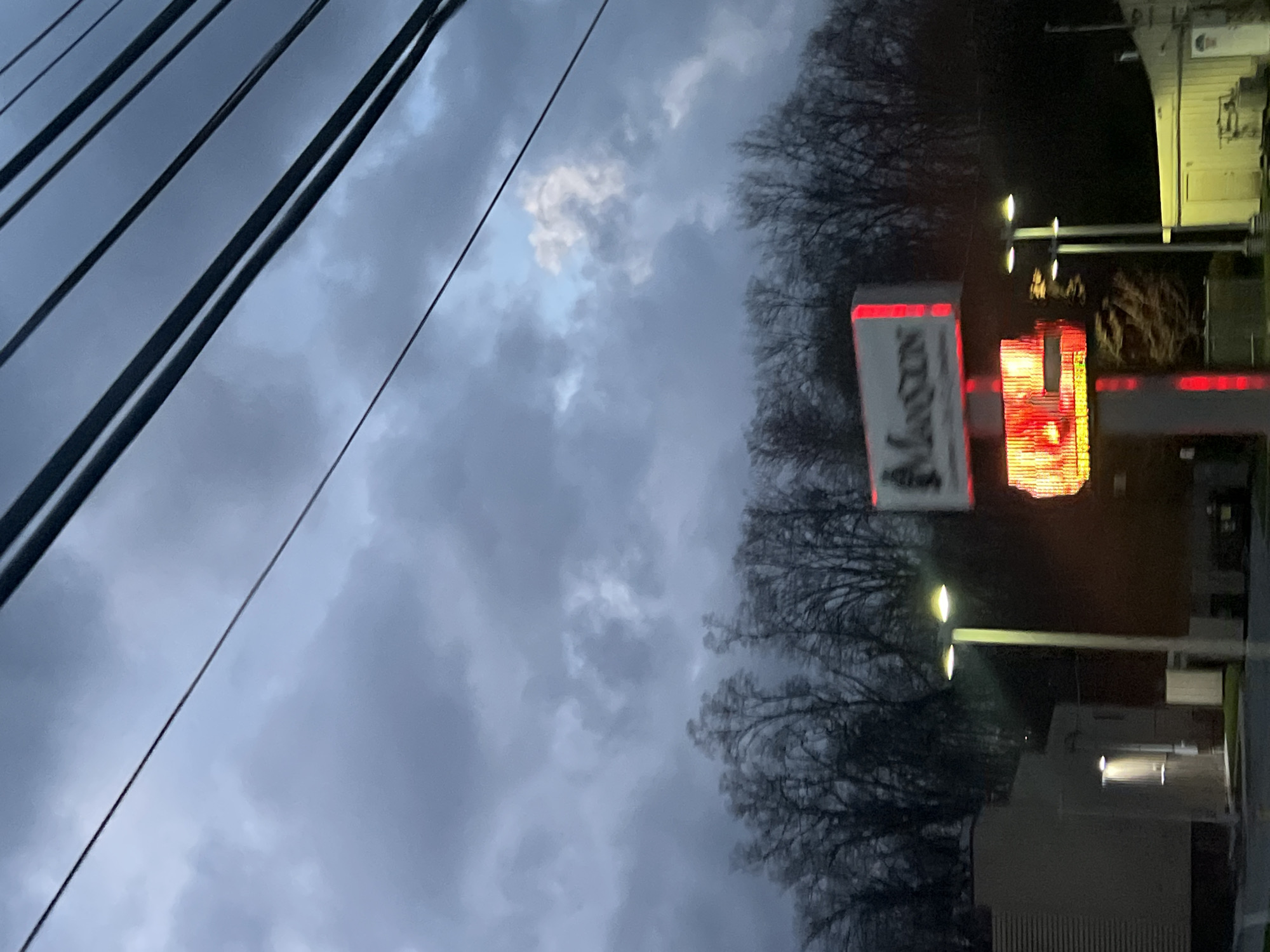 The image shows an outdoor view in Newburgh, United States at dusk or dawn. The sky is overcast with gray clouds. Power lines are visible in the upper left corner. A tree line separates the sky from buildings on the right. A sign that says "MAZON" in white with a red border is partly visible, along with an illuminated red sign nearby. Streetlights illuminate the buildings and a parking lot. The buildings appear to be low-rise structures.
