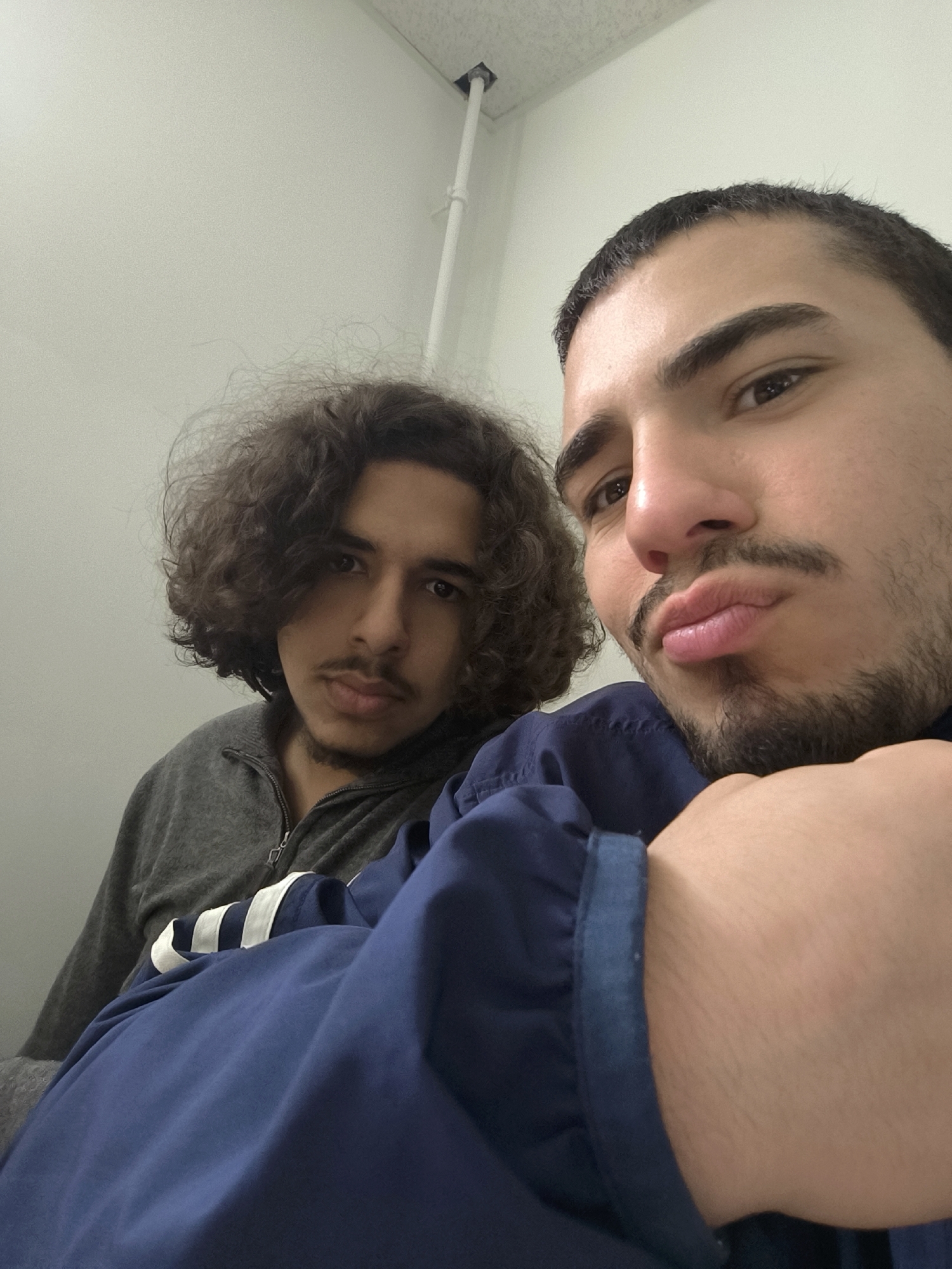 Two young men are in an indoor setting. One is in the foreground, wearing a navy blue collared shirt, posing for the photo with a pouting expression. The second man is in the background wearing a gray zip-up shirt. The background is a white wall with a white pipe affixed near the ceiling. The location is potentially in Schiltigheim, France.