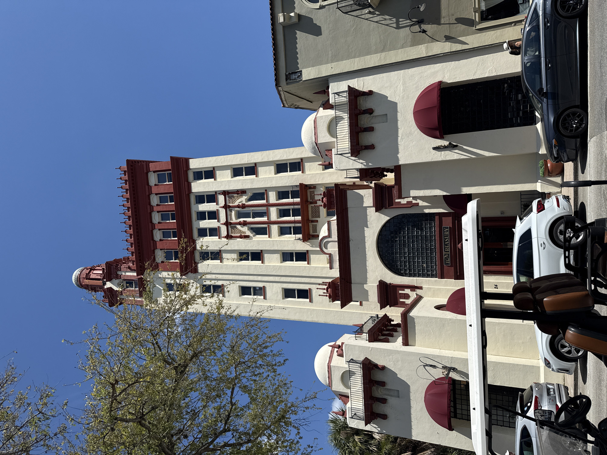 The image depicts the Treasury on the Plaza in St. Augustine, Florida, USA. The building is a multi-story structure with a white facade and red decorative elements. The architectural style includes domes and balconies. The sky is blue. A tree is visible in the foreground. Cars and a golf cart are parked on the street in front of the building.