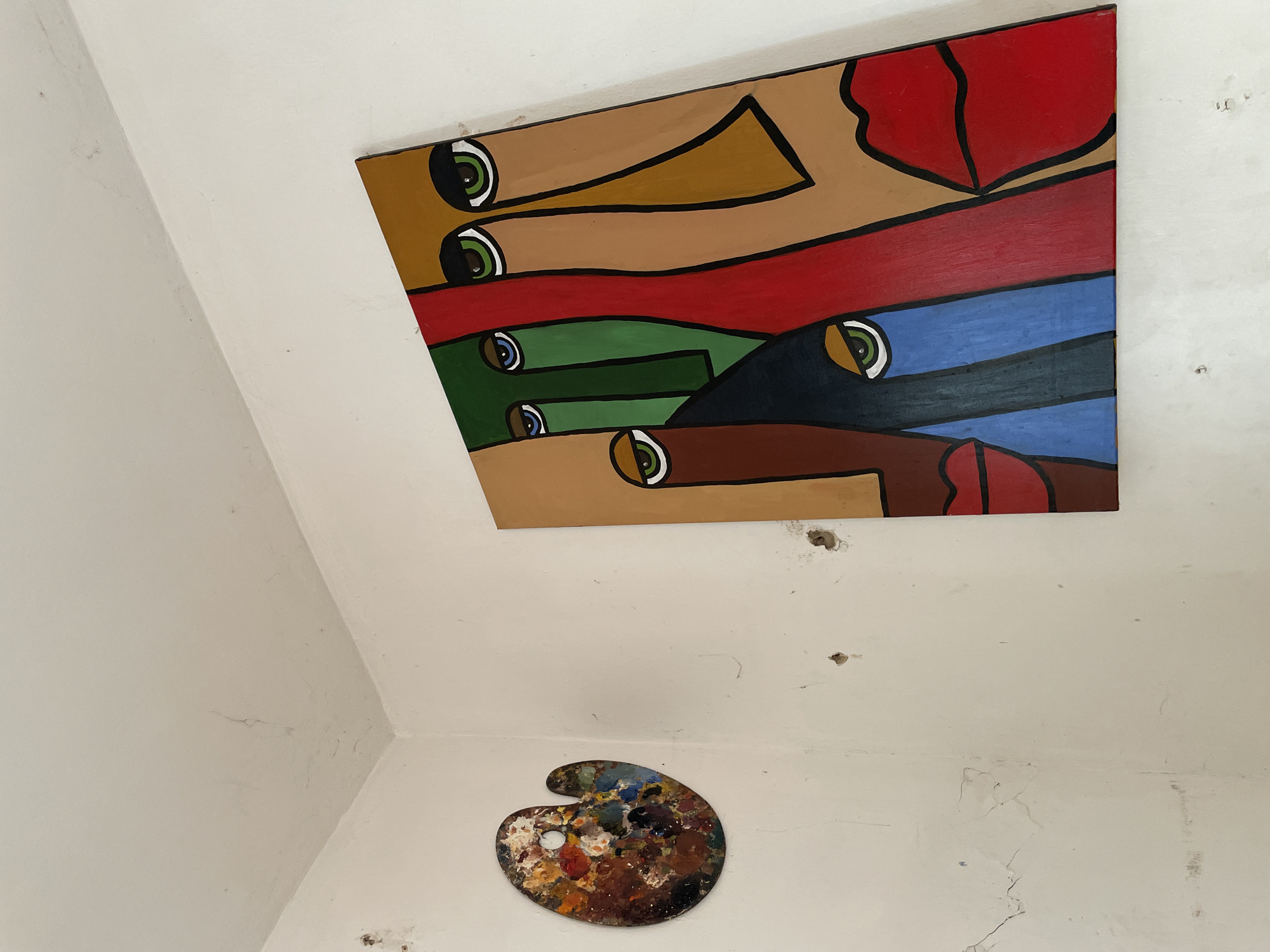 The media file captures an indoor scene, likely a room or hallway in Ado, Nigeria, featuring a prominent abstract painting and a painter's palette against off-white walls.
The main subject is a large, rectangular abstract painting mounted high on the wall, almost reaching the ceiling. The artwork employs a cubist or fragmented style, depicting multiple stylized faces or figures through overlapping and interlocking geometric shapes. These shapes are rendered in a vibrant palette of colors including tan, red, blue, green, and dark brown, all distinctly outlined in thick black lines. Numerous eyes with varied iris colors—green, blue, and brown—are scattered across the canvas, suggesting a multitude of gazes.
Below the painting, on a lower section of the wall, an artist's palette is displayed. It is kidney-shaped and heavily laden with a thick accumulation of dried paint in a diverse array of colors, including various shades of brown, blue, green, red, yellow, and a distinct white patch, indicating its regular use.
The surrounding walls and ceiling are painted an off-white color and show signs of age and wear. Visible details include hairline cracks, textured surfaces, and several small, dark holes or marks on the wall, particularly near the painting and above the palette. The overall setting suggests a lived-in space with an artistic presence. No discernible text is visible within the image.