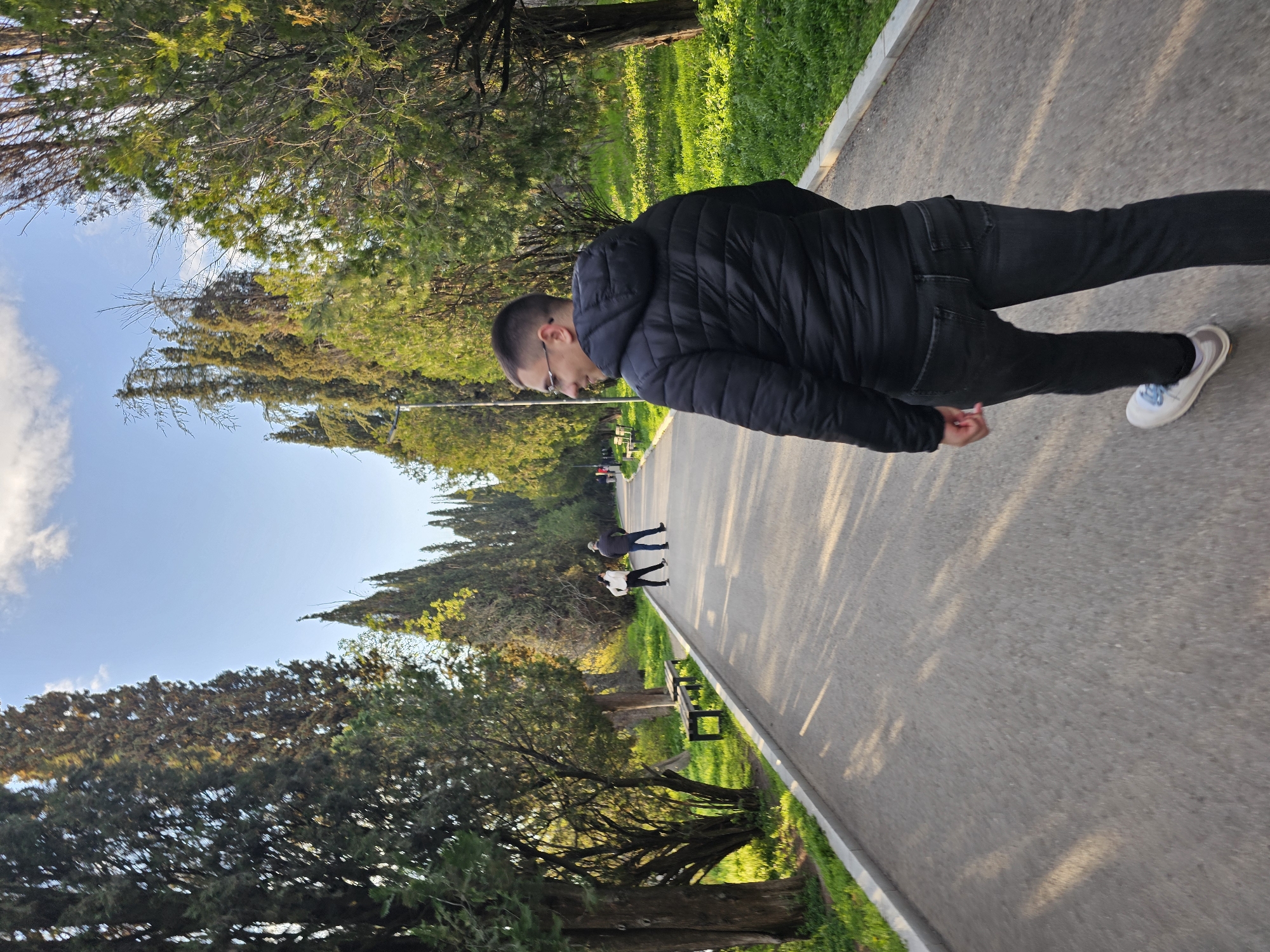 The image depicts a person walking on a paved path in a park-like setting in Stara Zagora, Bulgaria. The individual is wearing a black jacket, black pants, and white sneakers. They are facing the direction of travel along the path. The path is bordered by trees and green grass. In the distance, other people can be seen walking on the same path. The sky is blue with some clouds. The scene appears to be outdoors on a sunny day.