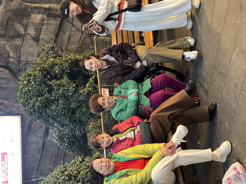 The image depicts five East Asian females in an outdoor public space within Yuzhong District, China. Four individuals are seated on a wooden slatted public bench with dark metal supports, while one stands to the right.
From left to right:
1. A female, appearing middle-aged or elderly, is seated, wearing a bright yellow puffer jacket over a red top, white pants, and white platform sneakers. She is smiling and looking directly at the camera. A partially visible white plastic bag with red text rests on the ground near her feet.
2. A female, appearing middle-aged or elderly, is seated next to the first, wearing a red puffer jacket over a patterned white sweater, brown wide-leg pants, and black low-heeled shoes. A black quilted handbag is on her lap. She is smiling and looking directly at the camera.
3. A female, appearing elderly, is seated, wearing a green button-up jacket, purple pants, and dark shoes. She has short, curly hair and a maroon crossbody bag. She is smiling and looking directly at the camera.
4. A female, appearing middle-aged, is seated, wearing a brown puffer jacket over a dark patterned top, olive-green wide-leg pants, and white sneakers. She is smiling and looking slightly to her right.
5. A female, appearing middle-aged, is standing to the right of the bench, wearing a white cropped puffer jacket with brown fur trim, a red long-sleeve top, white wide-leg pants, beige platform sneakers, and a black beret. She has a black crossbody bag and is holding a smartphone with both hands, looking at its screen, and smiling.
The background features a textured wall resembling rock formations, with dense green bushes and shrubs in front of it. Above the foliage, a white rectangular sign with red text and numbers (e.g., "C 工服", "6F", "SF") is visible. The ground is paved with dark gray rectangular stones. Lighting suggests late afternoon or early evening conditions.