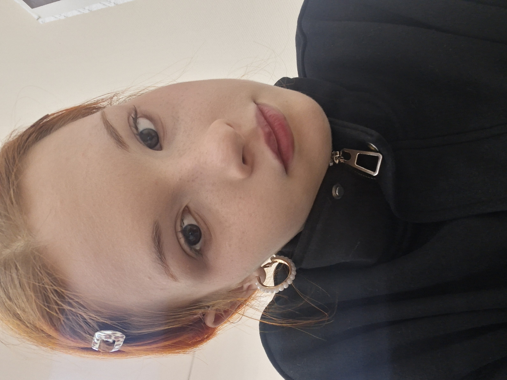 A close-up, rotated portrait features a young individual with fair skin, light reddish-brown hair, and light-colored eyes. The subject is looking directly at the camera with a neutral expression, enhanced by defined eyebrows and a reddish-pink lip color.
They are wearing a black jacket or coat, identifiable by a prominent silver-toned zipper pull on the right side of the frame. An earring in the right ear (viewer's left) is a gold-colored hoop adorned with small white beads or pearls. A clear, textured hair clip is visible in the hair on the left side of the frame (viewer's right).
The background is a plain, light-colored surface, suggesting an indoor setting. The image is taken from a low angle, with the subject's head tilted to the right, giving the appearance of a rotated perspective. The activity is a direct gaze portrait. No specific time of day or weather conditions can be determined from the visual cues. No text is visible within the image. The provided context for this scene is Chaikovsky, Russia.