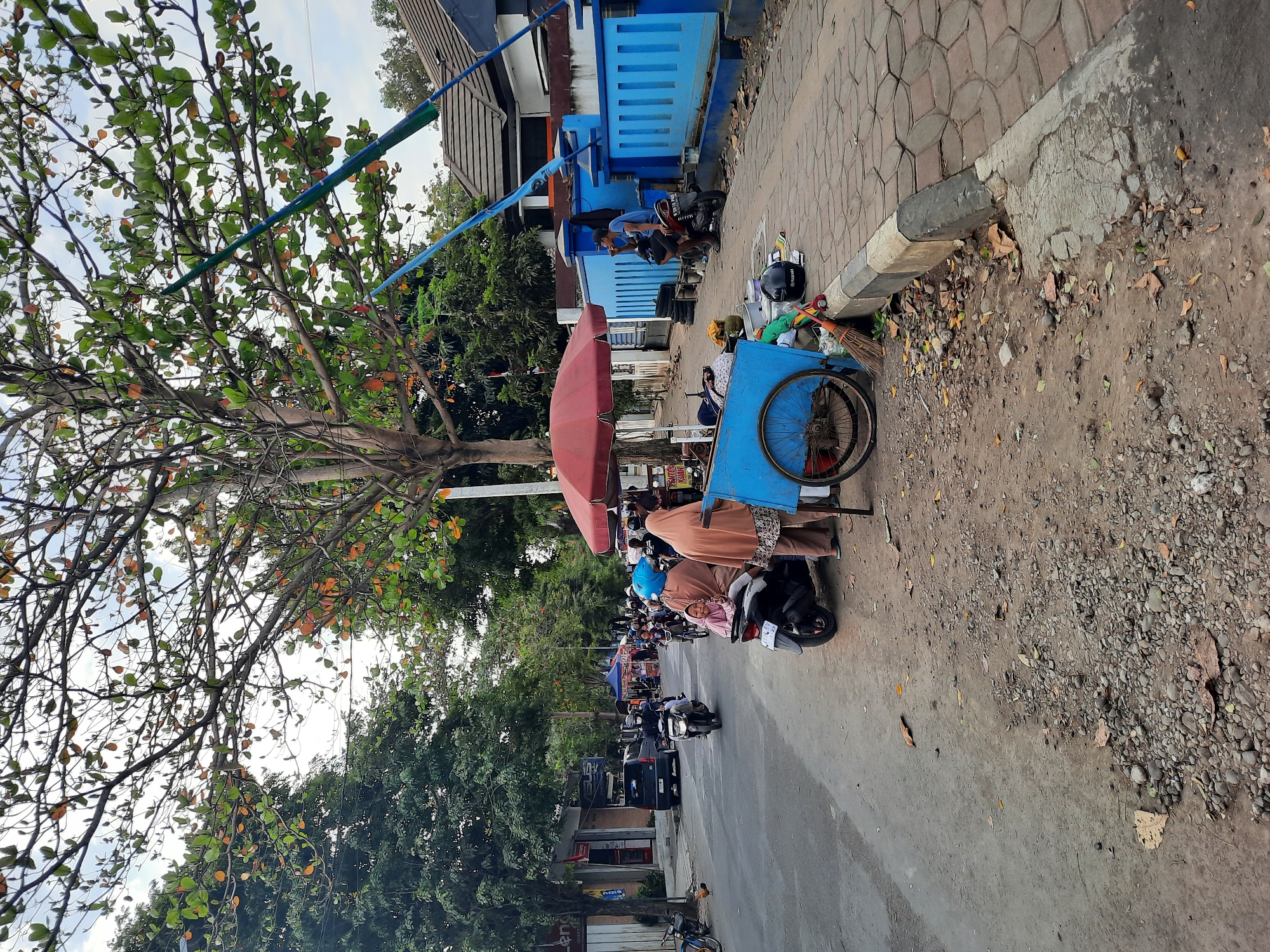 The image depicts a street scene in Kutoarjo, Indonesia. In the foreground, a blue pushcart is parked on a dirt shoulder next to a paved sidewalk. The pushcart has a red umbrella providing shade over what appears to be items for sale or storage. To the left of the pushcart, two people are seated on a motorcycle. One person, wearing a light brown headscarf and robe, is looking at the camera with a smile. Another person, wearing a blue headscarf, is seated behind them.
The street is moderately busy with traffic, primarily motorcycles. Several motorcycles are visible on the asphalt road, some with riders and passengers. On the left side of the street, there are buildings with storefronts, including one with signage that appears to read "Len". Trees with green and orange leaves line the street, casting dappled shade.
On the right side of the scene, a bright blue fence runs along the sidewalk. Behind the fence, a building with a brown tiled roof is visible. A man in a blue shirt is sitting on a motorcycle near the fence, appearing to be smoking. The sidewalk is paved with interlocking concrete blocks. The ground on the shoulder is a mixture of dirt, gravel, and fallen leaves. A broom and a helmet are placed near the pushcart. The overall atmosphere suggests a typical day in a bustling Indonesian town, with street vendors, local commerce, and daily life unfolding.