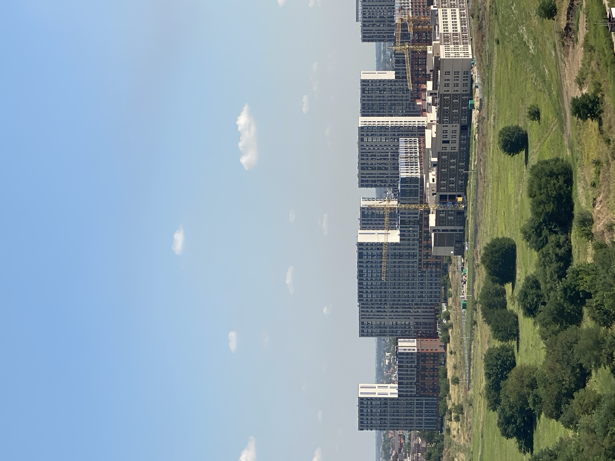 The image shows a panoramic view of a cityscape under a clear blue sky with a few scattered clouds. In the foreground, a grassy field with scattered trees and shrubs is visible. The middle ground is dominated by several multi-story buildings, including modern residential high-rises and a lower, longer building that appears to be a school or administrative complex. Construction cranes are present, indicating ongoing development. In the background, more buildings and a distant horizon are visible. The overall scene depicts a developing urban area with a mix of residential and possibly institutional structures, set against a backdrop of open green space.
