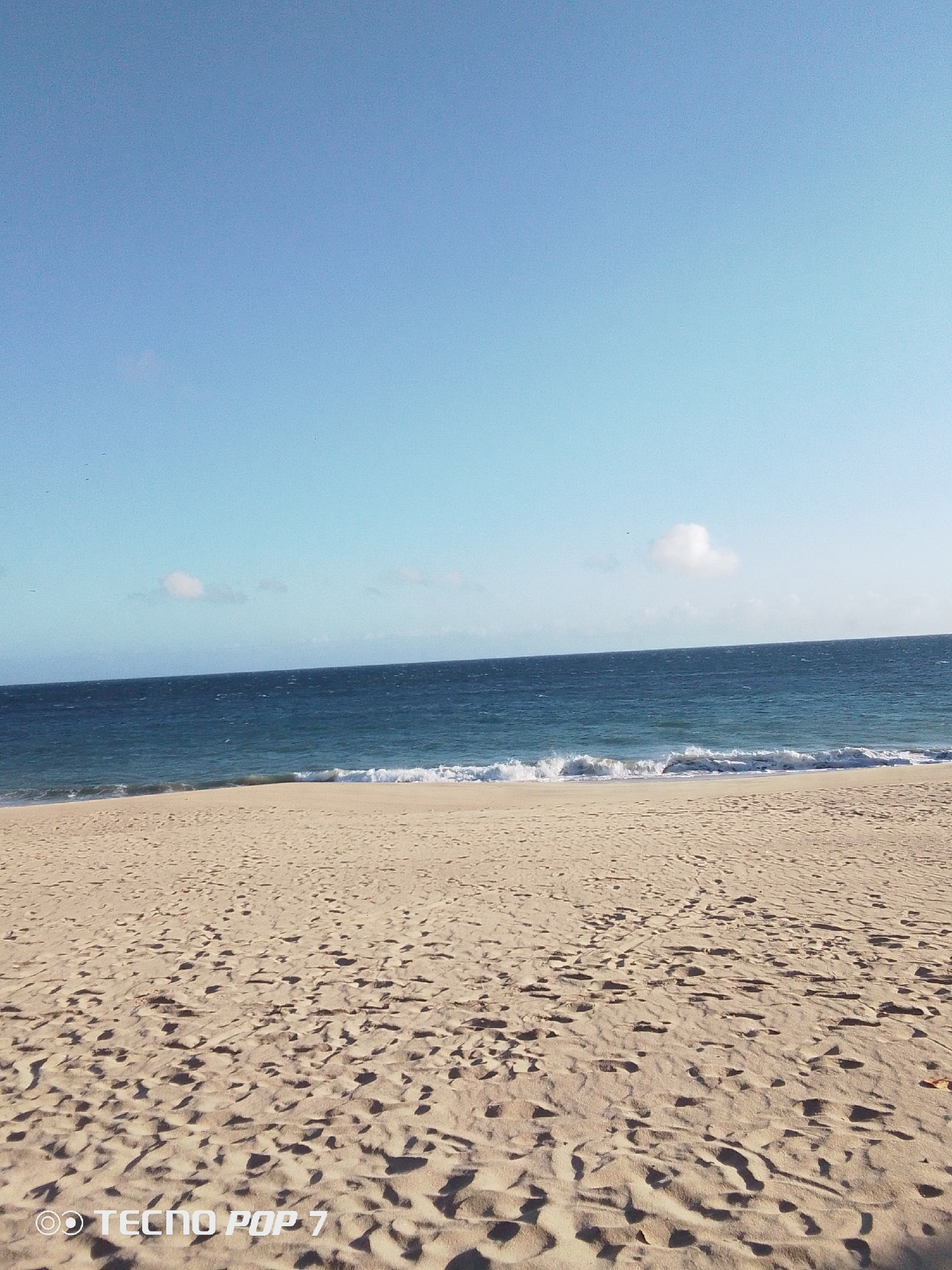 The image depicts a sunny day at a beach. The location is unspecified but likely in Pedro González, Venezuela, given context. The sky is a clear blue with scattered small clouds. The ocean is a darker blue with small waves breaking on the sandy shore. The beach is composed of light-colored sand, covered in footprints. The lower left corner of the image contains a watermark, "TECNO POP 7". No people are visible in the scene.