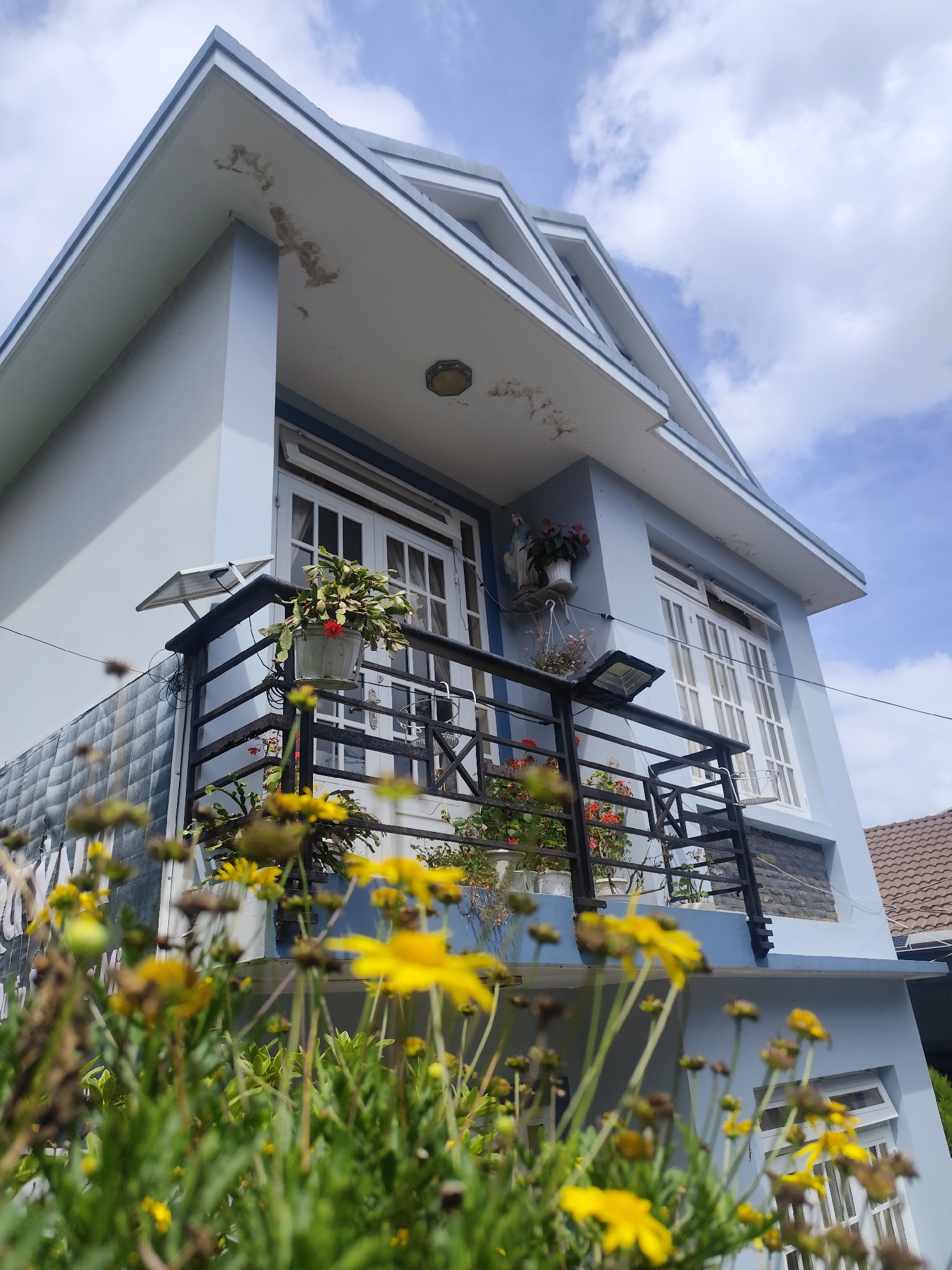 A low-angle outdoor shot captures the exterior of a light blue, two-story house with white-framed doors and windows, and a pitched roof with gray trim. The house is adorned with a black metal balcony on the second floor, featuring several potted plants, including one with red flowers and another with yellow flowers. Two solar-powered lights are mounted on the balcony railing. In the foreground, out-of-focus yellow flowers and green foliage dominate the lower portion of the image. The sky above is a mix of blue and white clouds. The image appears to be taken in Da Lat, Vietnam, a city known for its colonial architecture and abundant flowers.