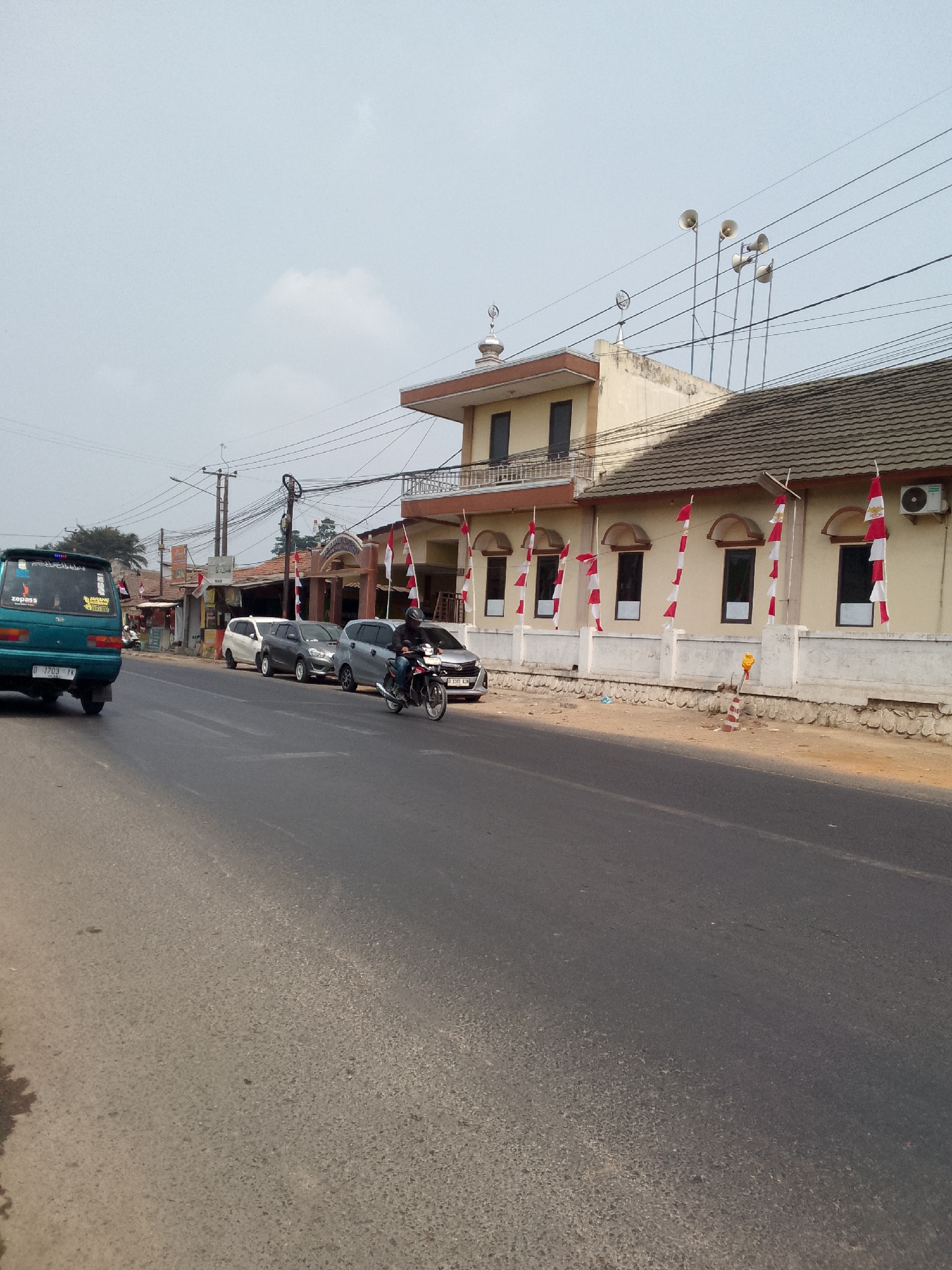 The image shows a street scene in Gunungmasigit, Indonesia, likely on a clear day given the bright lighting and lack of shadows. A paved road runs diagonally across the foreground and into the mid-ground. On the right side of the road, there is a light-colored building with red-tiled roofing. The building has several windows with arched tops, and it is decorated with red and white flags on poles. An air conditioning unit is visible on the exterior wall. A white concrete barrier runs along the front of the building.
On the left side of the road, a dark green minivan is parked. Further down the road, a motorcycle with a rider wearing a helmet is moving towards the right side of the frame. Several other cars are parked or driving on the road, including a white sedan and a gray sedan. In the background, there are more buildings, including one with multiple floors, and utility poles with many wires. Several loudspeakers are mounted on poles on top of one of the buildings. The overall environment appears to be a suburban or semi-urban area with a mix of residential and commercial structures.