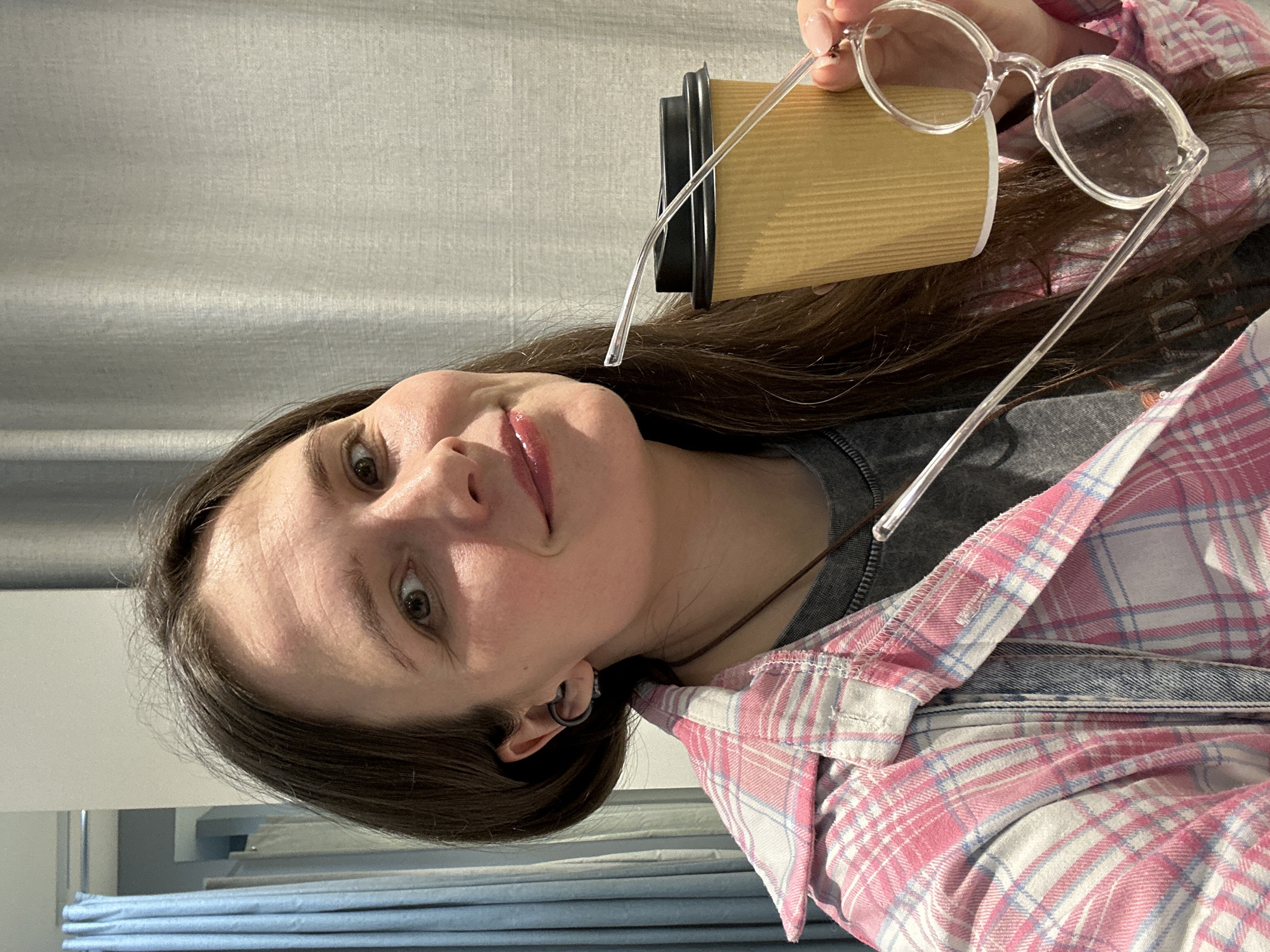 A young woman is captured in a casual, portrait-oriented selfie, looking directly at the camera with a soft smile. She has long, dark brown hair, partially visible on the right side of the frame. In her right hand, she holds a light brown, corrugated disposable coffee cup and a pair of clear-framed round glasses, both positioned near her head. Her left eye is clearly visible, and she wears a subtle pink lip gloss. A small dark hoop earring is visible in her left ear.
She is dressed in a grey t-shirt layered under an open flannel shirt featuring a pink, white, and light blue plaid pattern. A hint of denim fabric can be seen beneath the flannel on her right side.
The background consists of a light grey, textured curtain or wall covering in the upper portion. Below this, a white surface is visible, and further down, some folded light blue textiles or fabric are stacked. The lighting is soft and even, suggesting an indoor setting, likely during the daytime. The overall impression is relaxed and pleasant. There is no visible text in the image. The photo was taken in Šušanj, Montenegro.