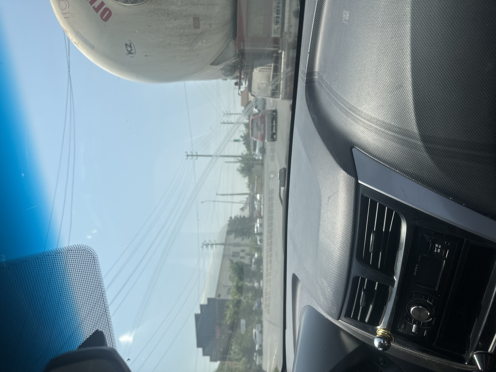 The image is taken from the interior of a vehicle, looking out through the windshield. The dashboard of the car is visible in the foreground, featuring a textured surface and an audio system. Outside, the scene appears to be a city street with moderate traffic under a bright, clear sky.
Several cars are on the road, and a large tanker truck is prominent on the right side of the frame. The tanker has some lettering visible, including "OT" and "KZ". In the background, buildings, trees, and utility poles line the street. Power lines crisscross the sky. The overall environment suggests a sunny day in an urban or semi-urban setting. Based on the presence of the tanker truck with "KZ" on it, the location is likely Manas, Kyrgyzstan. There are no people clearly visible in the image.