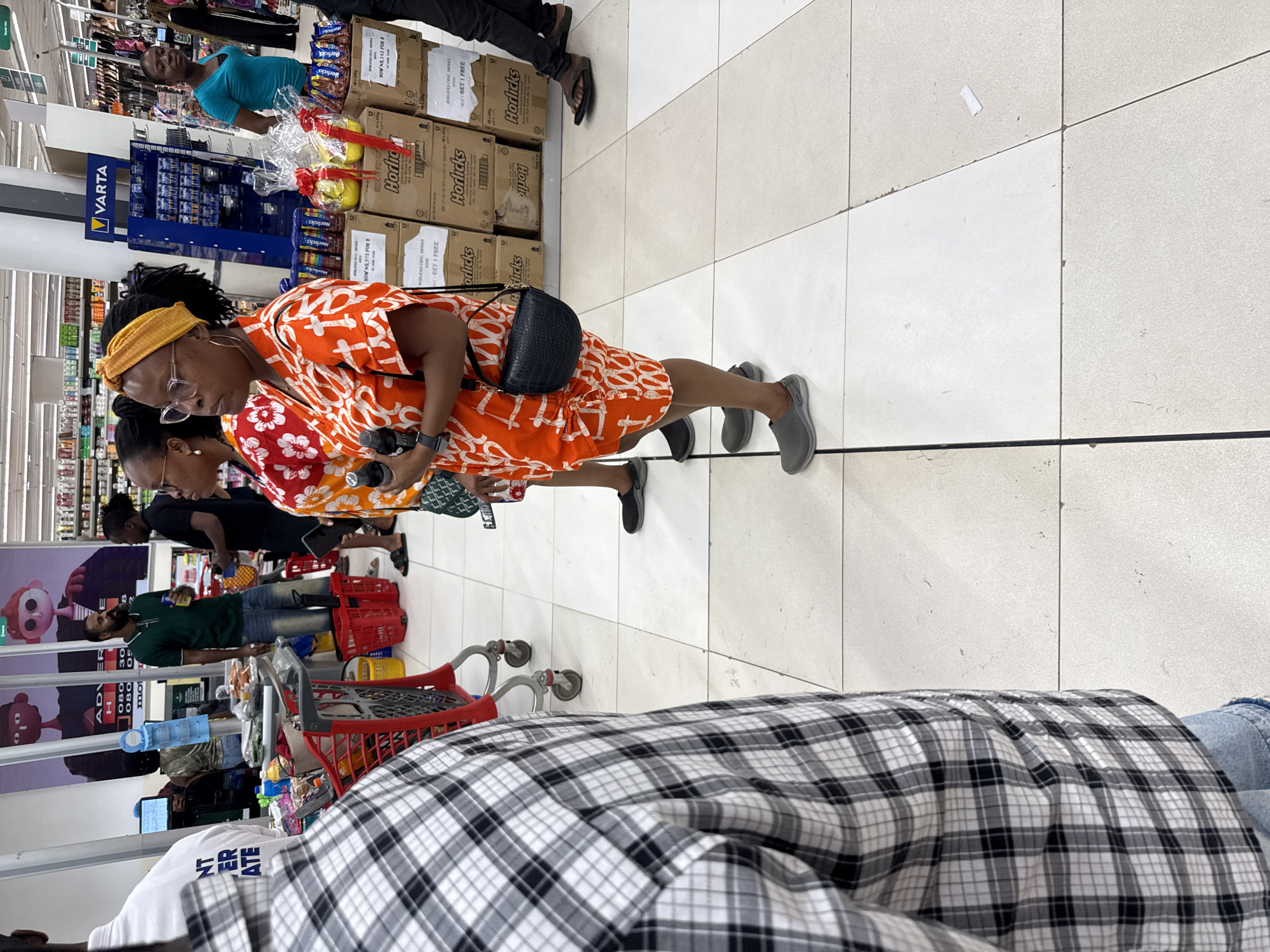 The image shows a shopping scene inside a supermarket in Abuja, Nigeria. There are multiple shoppers, including a woman in an orange and white patterned dress with a yellow headband and eyeglasses who appears to be browsing items. Another woman is standing beside her. There are shopping carts and shelves filled with various products, including boxes of Horlicks and Varta batteries. One person is wearing a plaid shirt. The floor is tiled.