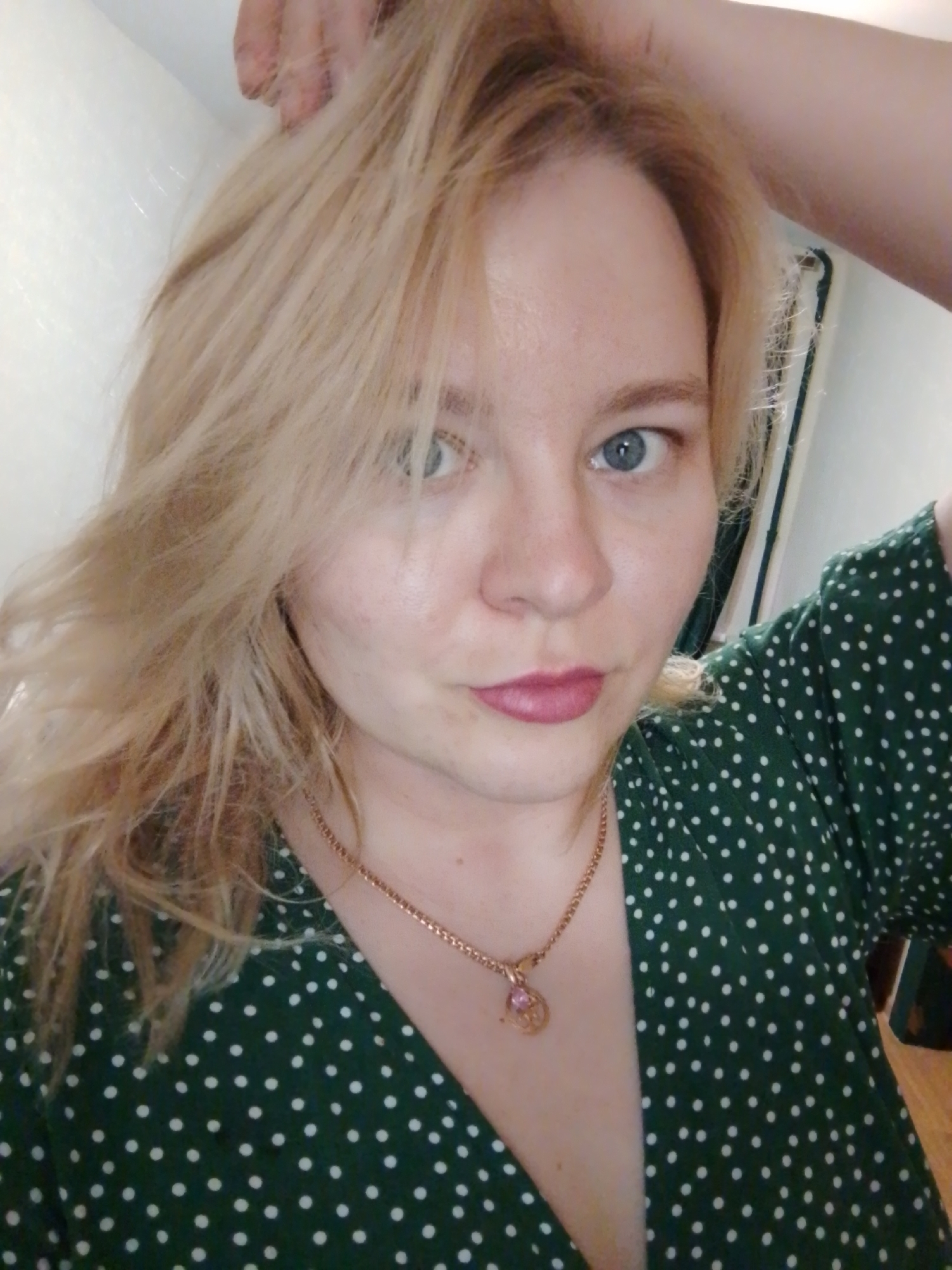A selfie of a woman with blonde hair and blue eyes. She is wearing a green dress with white polka dots and a gold necklace with a pink pendant. Her right hand is raised to her hair. The background is a plain white wall. The location is not discernible from the image.