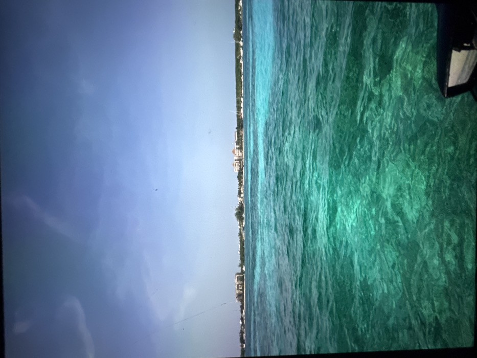 A coastal scene displays a clear blue sky meeting the turquoise ocean. Buildings line the horizon, suggesting a populated beachfront. The water is textured, indicative of gentle waves, and reveals varying depths through different shades of green. A corner of an object, potentially a boat or structure, is visible in the upper right.
