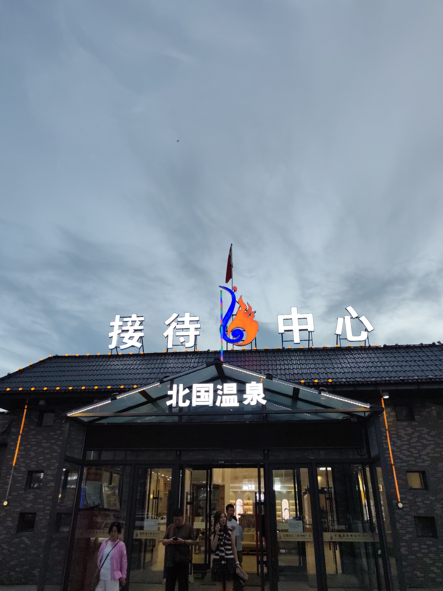 The image shows the exterior of a building with Chinese characters on a prominent sign. The sign, illuminated with white lights, reads "接待中心" (Reception Center) and above it, "北国温泉" (Northern Country Hot Spring). A stylized orange and blue flame graphic is placed between the two parts of the sign. The building has a dark, tiled roof with small orange lights along the eaves. A glass entrance with doors is visible at the bottom of the building. Several people are standing near the entrance, with some walking in and out of the building. The sky above is overcast with clouds, suggesting it might be late afternoon or early evening. The building appears to be located in the city of Lindian, China, as indicated by the text on the signage.