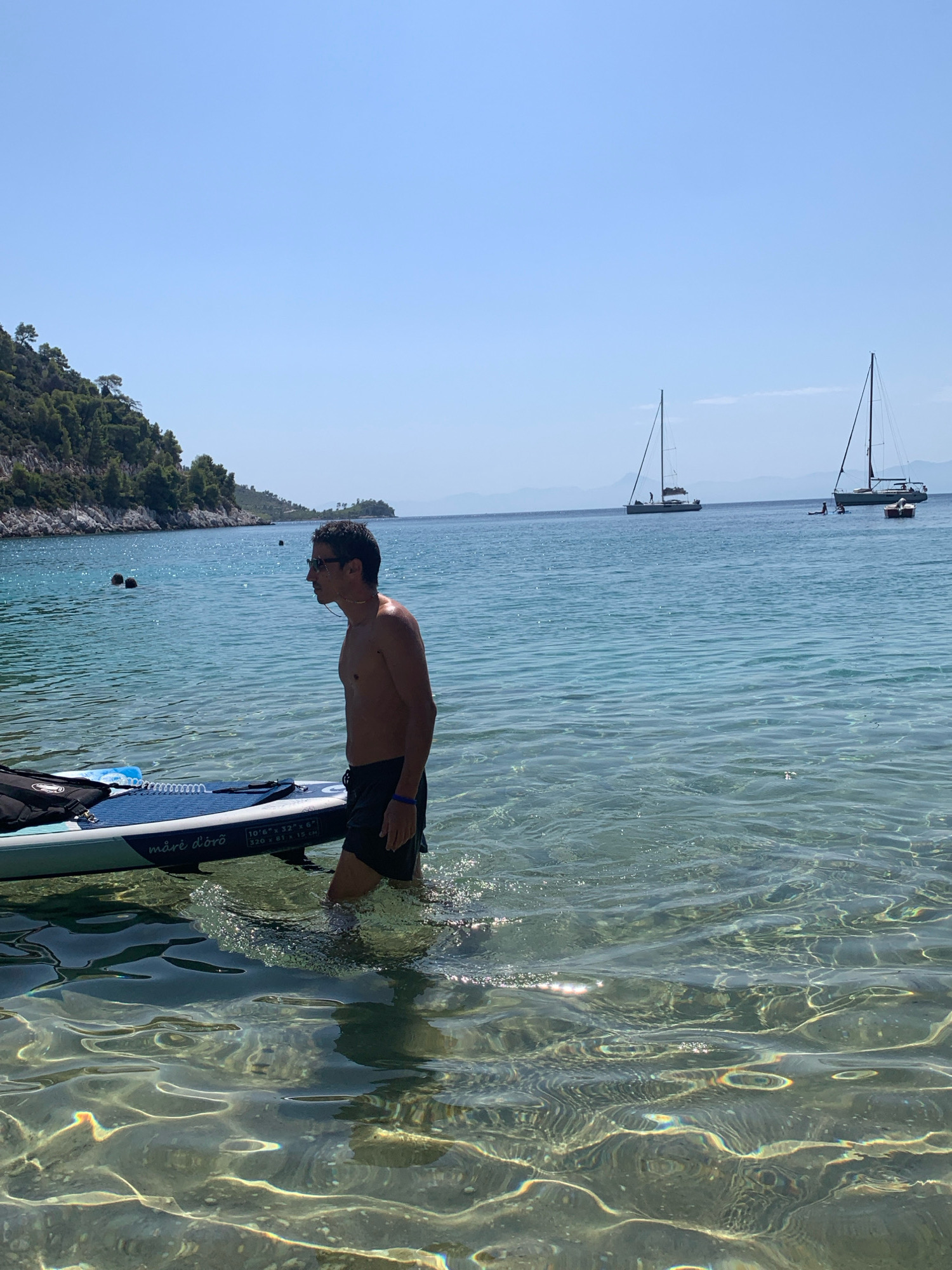 The image shows a man standing in the clear, shallow water of a beach in Aliki, Greece. He is shirtless, wearing dark shorts and sunglasses. To his left, a stand-up paddleboard is partially submerged in the water, with a black bag resting on it. The water is crystal clear, revealing the sandy bottom. In the background, a lush green, tree-covered hillside slopes down to the water's edge. Further out in the sea, two sailboats with tall masts are visible, along with a smaller boat. The sky is a clear, bright blue. The sun is shining, creating reflections and ripples on the water's surface. Two other individuals are partially visible swimming in the distance near the shore.