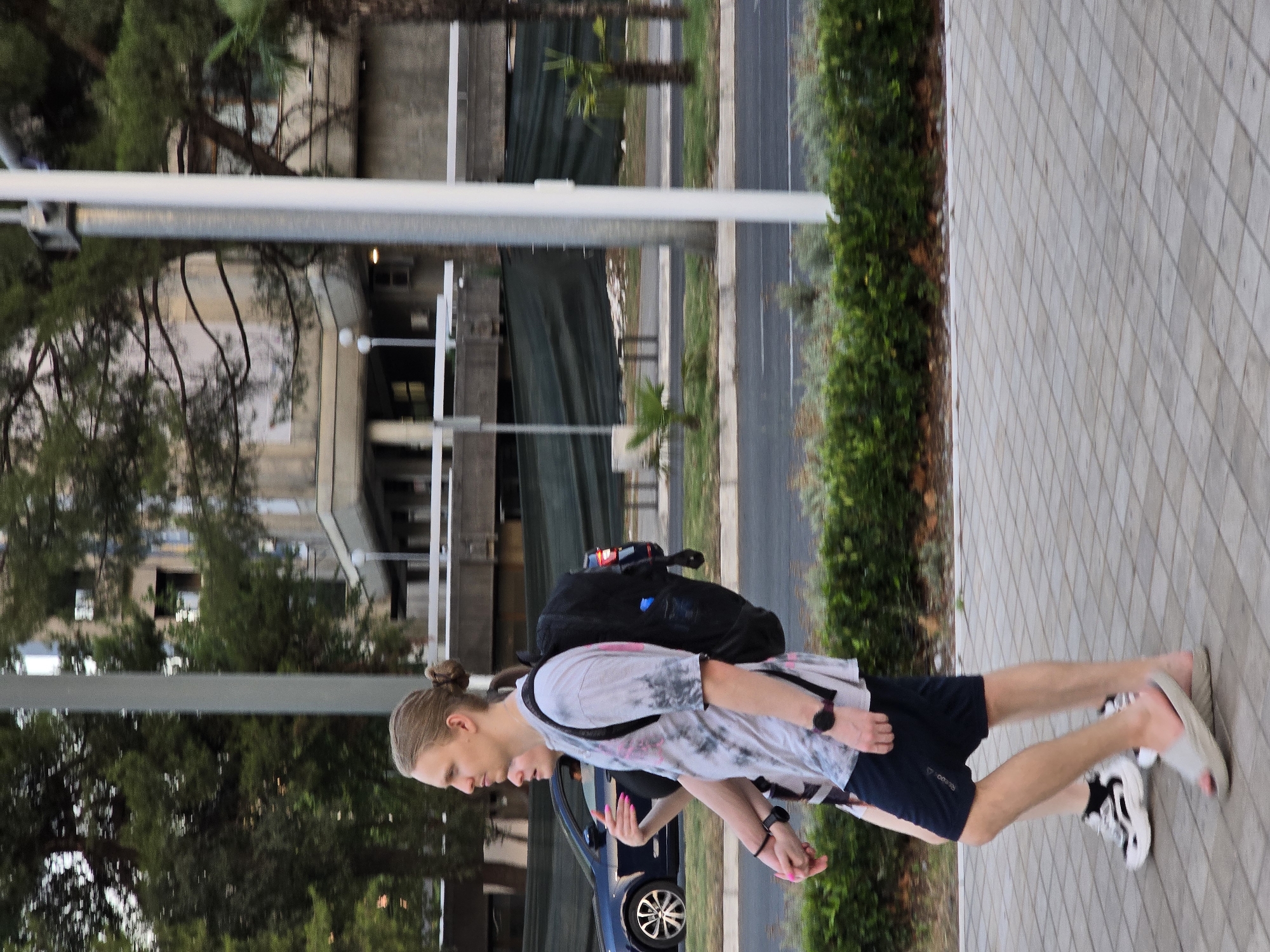 In Podgorica, Montenegro, a young Caucasian man with long blonde hair pulled into a bun and a woman with pink fingernails are walking hand-in-hand on a paved pedestrian walkway. The man is wearing a tie-dye t-shirt, dark blue shorts, and light grey slides. He is carrying a black backpack. The woman is wearing a black garment. They are walking towards the left of the frame. A dark blue sedan is visible in the background, driving on a road to their left. The environment includes green foliage, trees, and a building with a concrete facade. A tall white pole is positioned on the right side of the frame.
