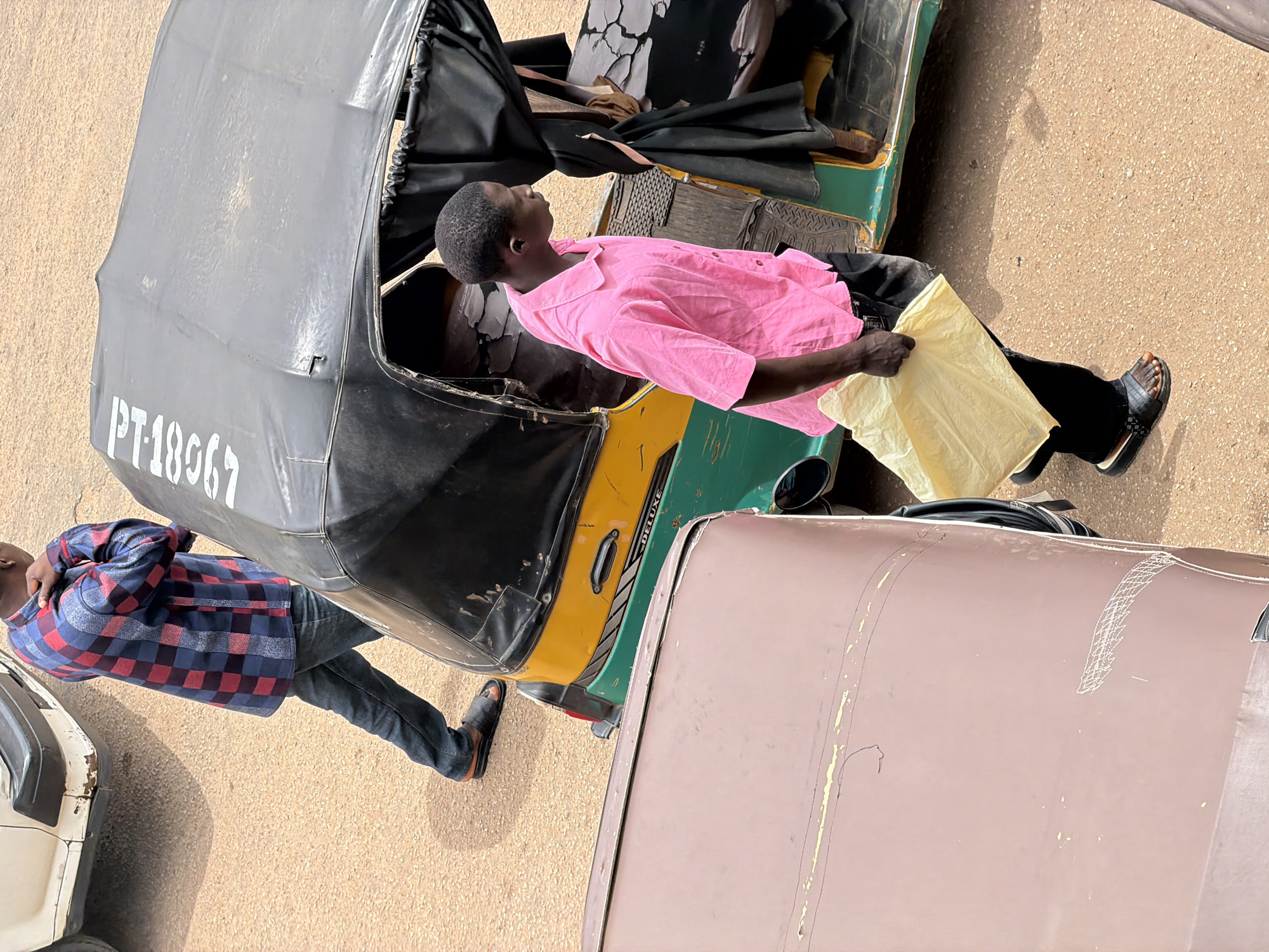 In Bukuru, Nigeria, two men are seen from a high angle. The primary subject is a driver of a three-wheeled auto-rickshaw, a common mode of transport. He is leaning back in his seat, wearing a bright pink shirt and black trousers, with a yellow cloth resting on his lap. The auto-rickshaw itself is black with a green and yellow body. Visible text on the vehicle reads "PT-18967".
To the left of the auto-rickshaw, another man is standing, looking down. He is wearing a blue and red checkered shirt and dark trousers. He appears to be in motion, with one leg bent and his hand on his hip, possibly disembarking or having just disembarked from another vehicle.
The ground beneath them is a sandy, gravelly surface, typical of outdoor environments in many parts of Nigeria. The lighting suggests it is daytime, with shadows cast on the ground, indicating a bright sun. The overall scene depicts a moment of rest or transition in a busy transportation hub or street. There are no explicit indicators of weather or strong emotions, but the casual poses suggest a normal, everyday situation. The presence of another vehicle partially visible on the left and a large, brown, possibly parked vehicle on the right frames the auto-rickshaw and its occupants.