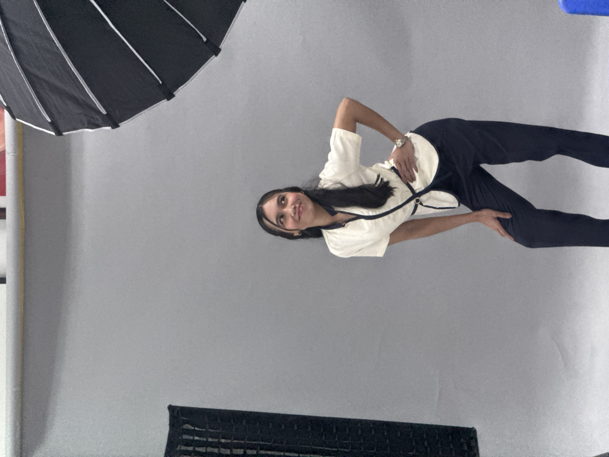 In a studio environment with a plain grey backdrop, a young woman of Asian descent stands posing. She has long, dark hair and is wearing a white top with dark trim and dark pants. Her left hand is on her hip, and her right hand is resting on her bent left knee. To the left of the frame, a black softbox is visible, and to the right, a portion of a large octagonal umbrella lighting modifier is present. The lighting suggests a professional photoshoot is taking place. There is no indication of any specific location within Davao City, Philippines.