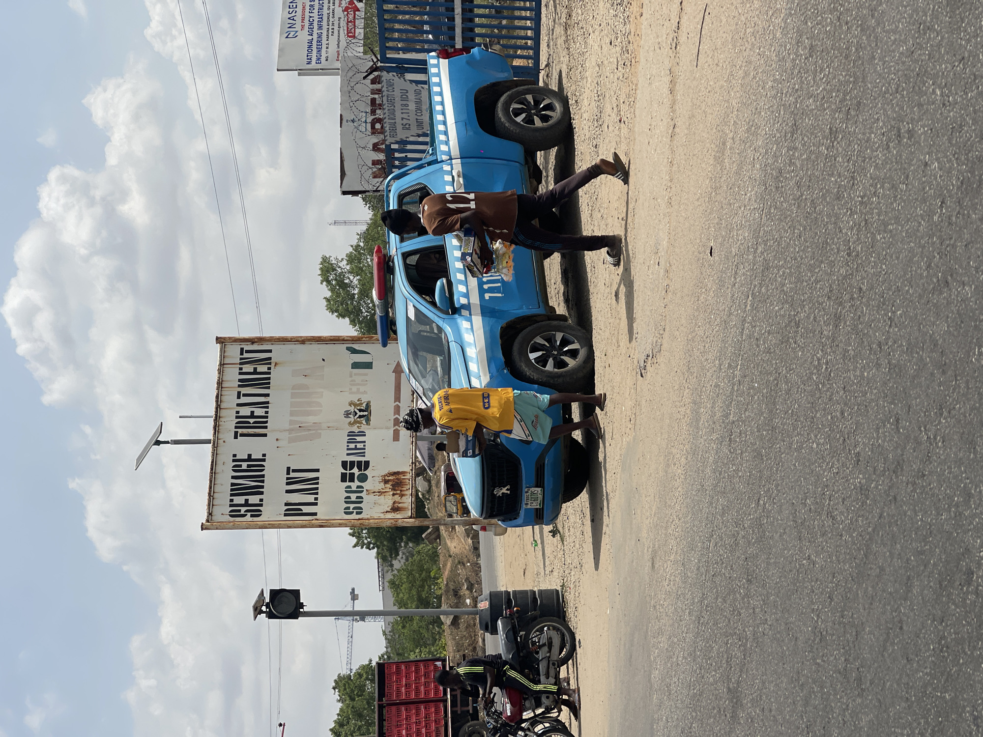 This image, taken in Karmajiji, Nigeria, captures a vibrant roadside scene under a bright, partly cloudy sky. A light blue pickup truck, identified as belonging to the "FEDERAL ROAD SAFETY CORPS" with "RS 7.118 IDU," "UNIT COMMAND," and "111" on its side, is parked on the dirt shoulder. Its license plate reads "C-607RS."
Two men are interacting with the truck. One, wearing a brown shirt with "12" on the back and dark pants, is at the driver's side window, holding a tray of items, suggesting a roadside vendor making a sale. Another man, in a yellow shirt with "TECATE," "AFIRME," and "H-E-B" printed on it, and light blue shorts, stands near the front passenger side of the truck.
To the left, a large rusty sign reads "SEWAGE TREATMENT PLANT," "WODA," and "SCCM AEPB." Below it are a traffic light and two motorcycles, one red with a black seat, loaded with red plastic crates. A person in a black and yellow striped shirt sits on the red motorcycle.
In the background, behind the blue truck, a white sign prominently displays "NASENI," "THE PRESIDENCY," and "NATIONAL AGENCY FOR SCIENTIFIC ENGINEERING INFRASTRUCTURE," along with an address in Abuja and an email contact. The road is asphalt, bordered by a dirt shoulder. The scene appears to be an ordinary daytime street view, potentially showing commerce or official activity.