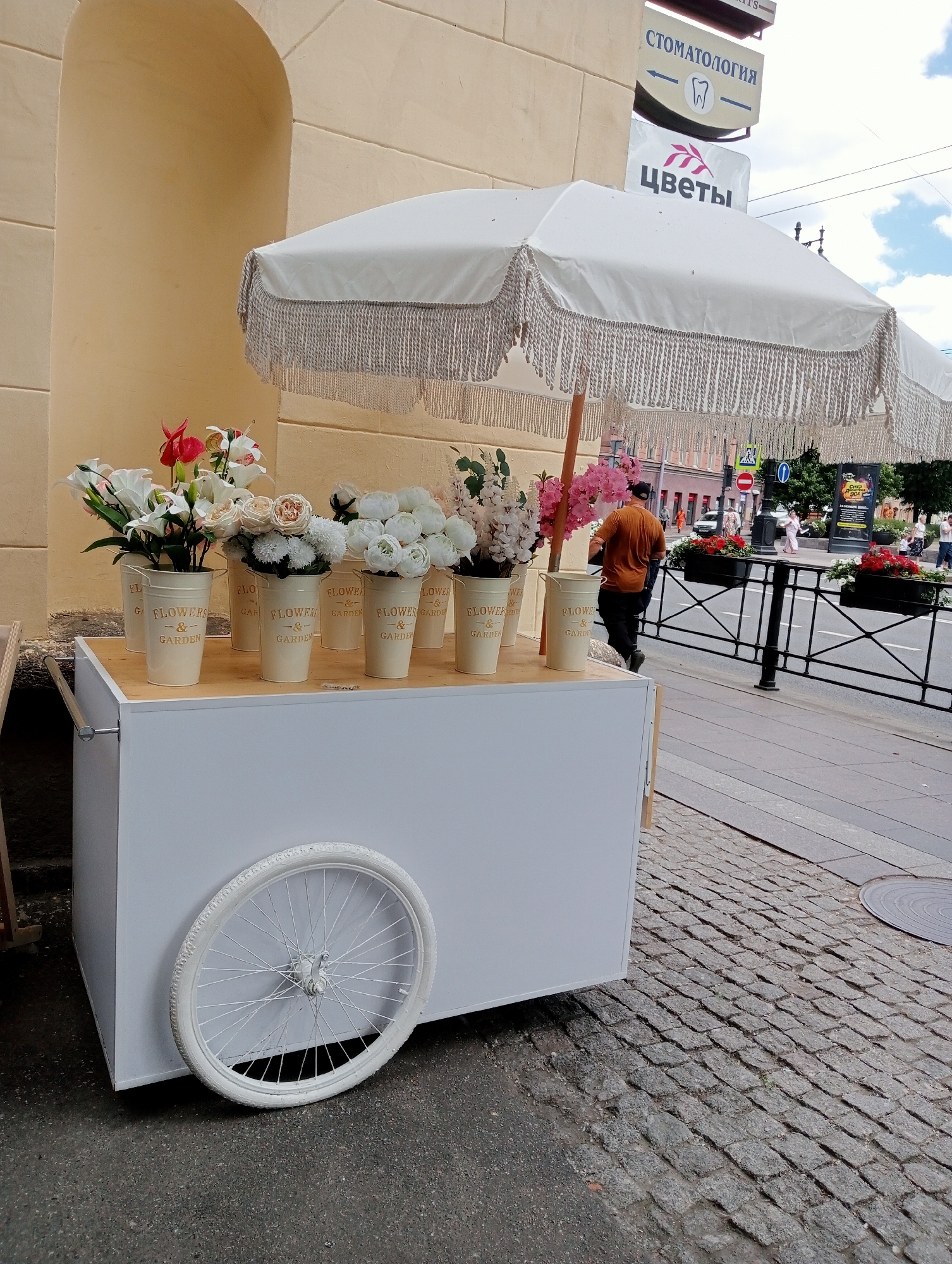 This is a photograph taken outdoors in Saint Petersburg, Russia. In the foreground, a white flower cart with a white bicycle wheel is parked on a cobblestone sidewalk. The cart is laden with several buckets of artificial flowers in various shades of white, pink, and red. A white umbrella with fringed trim is extended over the cart. In the background, a man in an orange shirt and black pants walks away from the camera, facing a street with buildings and pedestrians. Signs with Russian text are visible on the building to the right, including one that reads "СТОМАТОЛОГИЯ" (Dentistry) and another with the word "ЦВЕТЫ" (Flowers). The scene appears to be a street-side flower vendor.