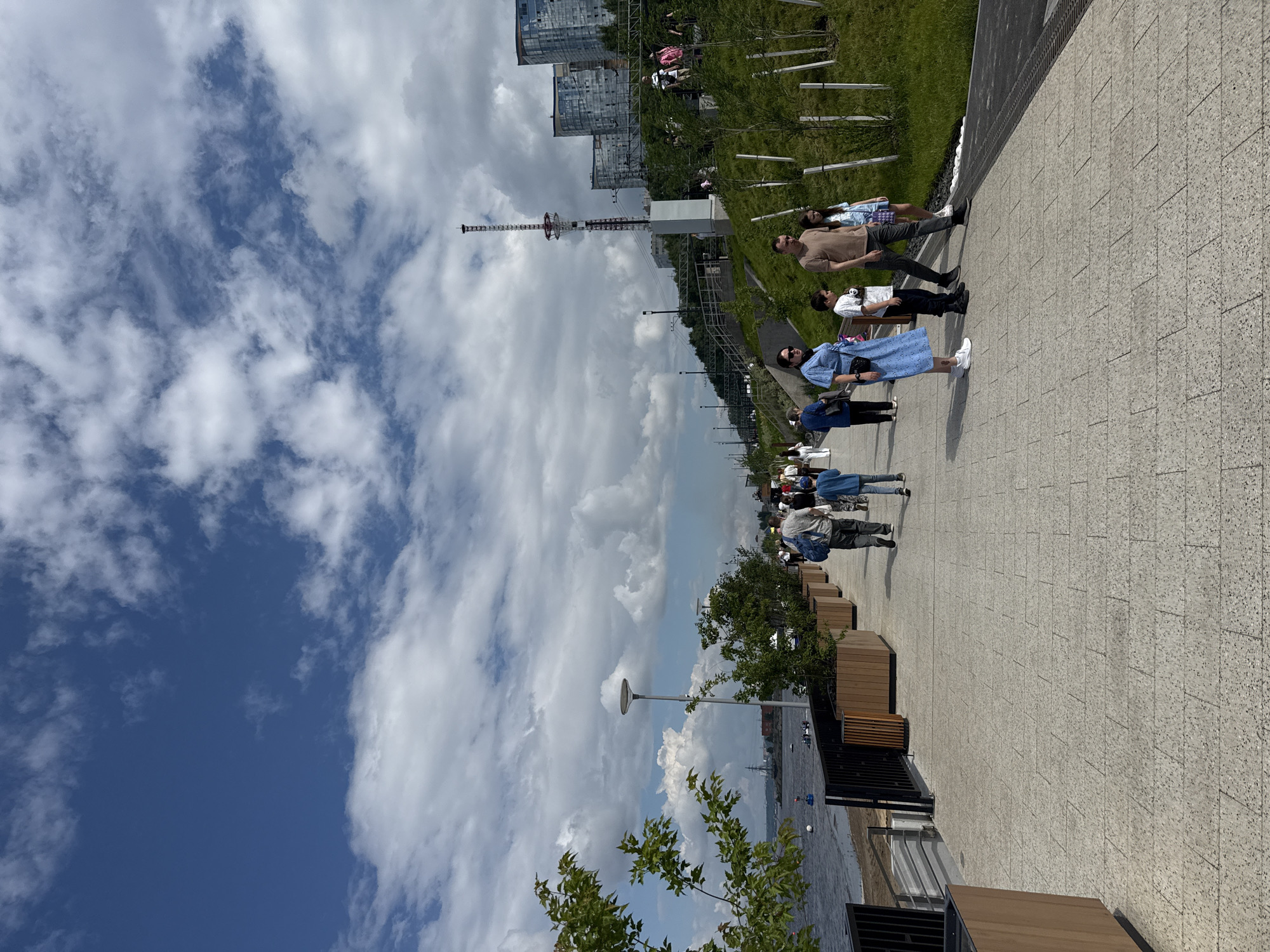 This image depicts a view of the Perm embankment in the city of Perm, Russia. The foreground shows a paved walkway lined with modern planters and greenery. Several people are seen walking along the embankment, some individually and others in groups, enjoying the day. In the middle ground, the Kama River is visible on the left, with some boats on the water. The background features a cityscape with tall, modern buildings and a distinctive radio tower. The sky is partly cloudy, with a mix of blue sky and white clouds. The overall scene is one of a vibrant urban public space on a pleasant day.