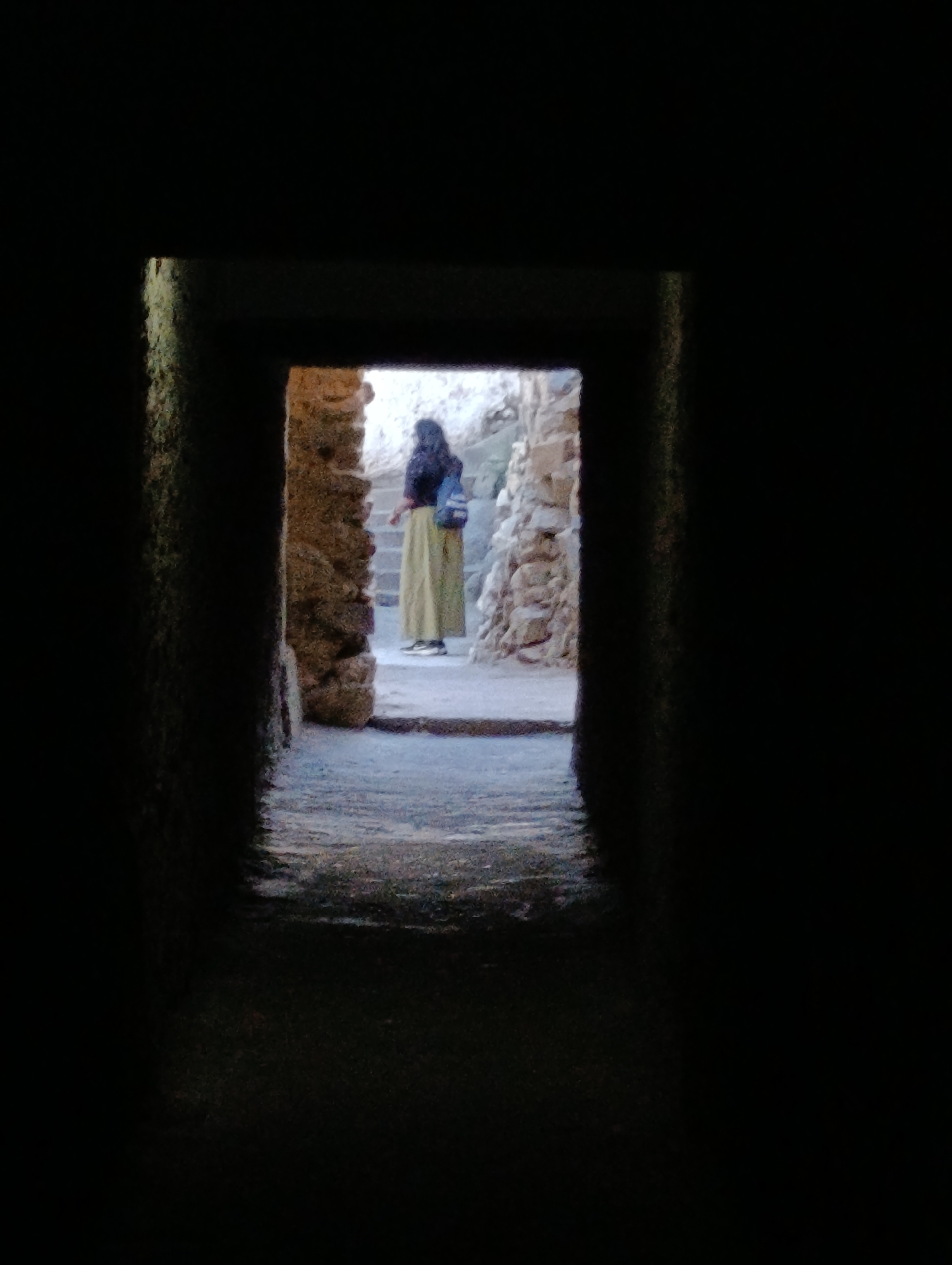A full-body, outdoor, eye-level shot captures a person from the chest up, standing within a rectangular stone archway. The person, facing away from the viewer, is wearing a dark top, a light-colored, floor-length skirt, a light-colored backpack, and dark shoes. They are positioned on a stone pathway that leads to a staircase on the right. The archway's rough stone walls are visible on the left and right, framing the scene. The lighting is dim, suggesting an indoor or shaded environment. The background behind the person appears to be more stone structures and a dimly lit pathway.