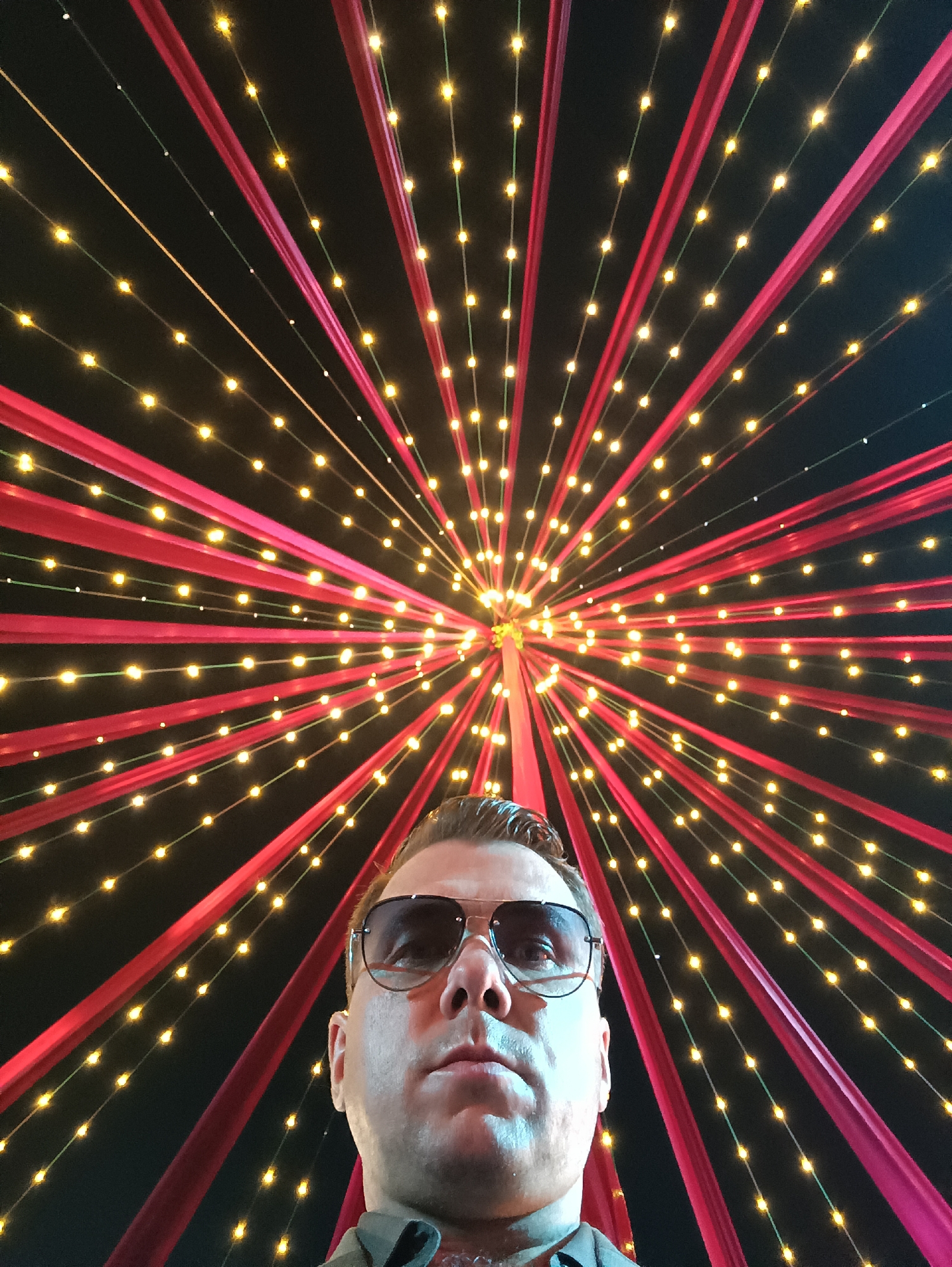 A low-angle shot shows a man wearing sunglasses. The man is fair-skinned, and his face is well-lit. The background is a burst of red cloth strips emanating from a central point and interspersed with strings of small, warm-toned lights. The location appears to be Kalamassery, India.