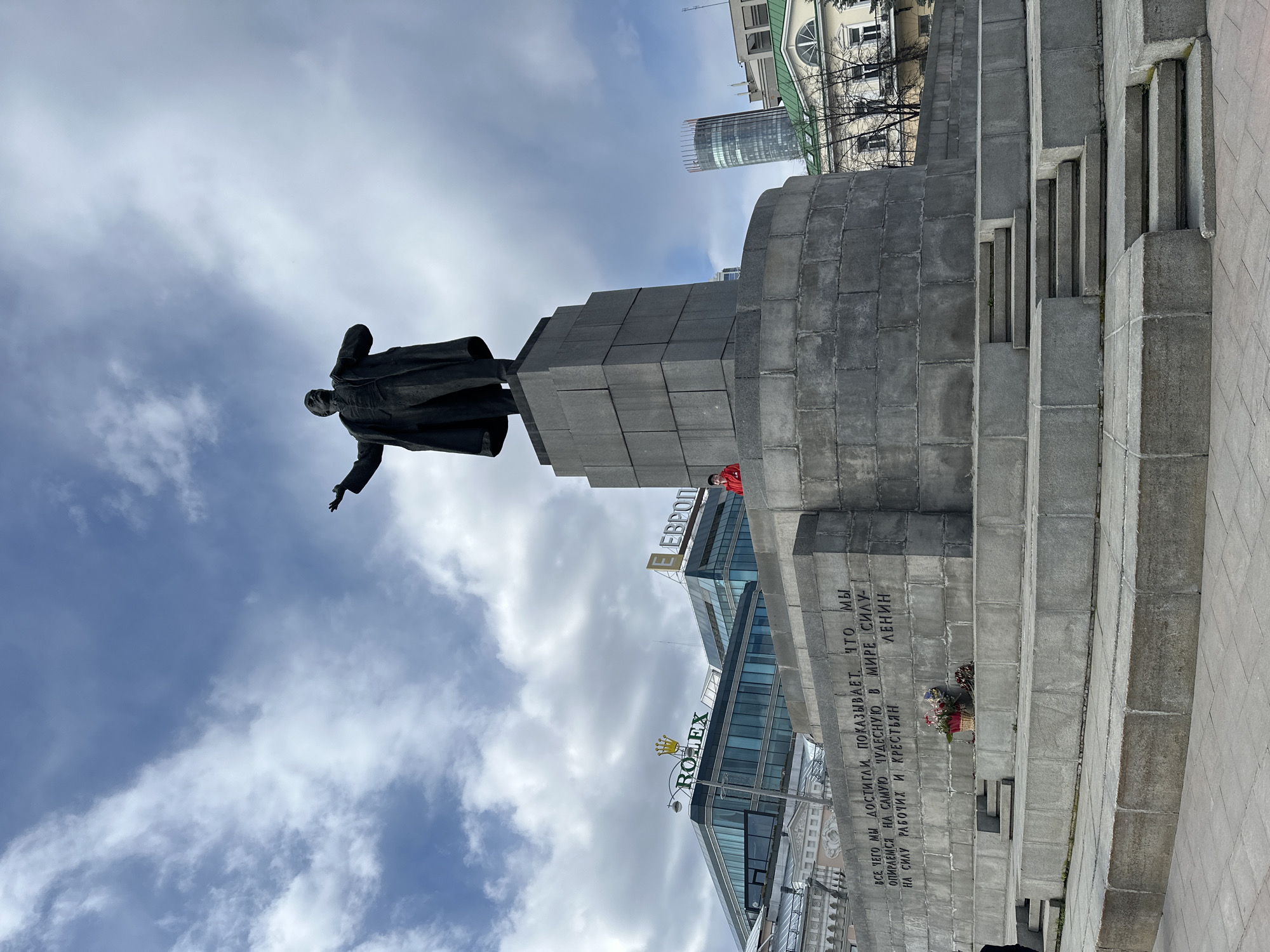 The image depicts the statue of Vladimir Lenin in Yekaterinburg, Russia. The statue is situated on a tiered stone plinth. In the background, modern buildings are visible, including one with Rolex and Evropa signage. A memorial wreath lies at the foot of the monument, near engraved Cyrillic text on the stone. The sky above is partly cloudy.