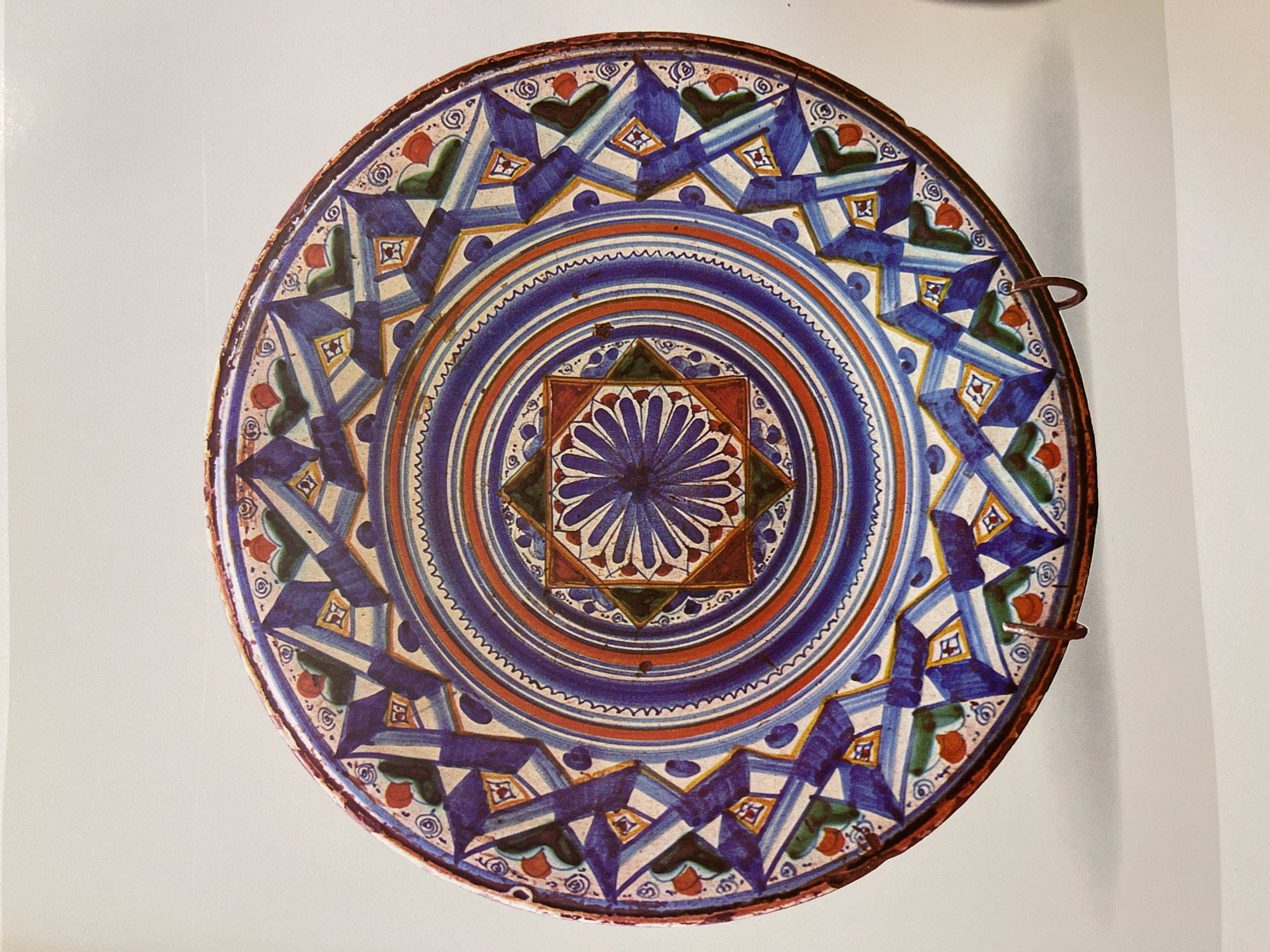 The image depicts a colorful, hand-painted ceramic plate with intricate geometric and floral designs. The central design features a stylized flower or rosette within a square frame. Concentric circles of blue and orange radiate outwards from the center. The outer ring displays a pattern of alternating blue and white geometric shapes, interspersed with small green and red elements. Two small metal loops protrude from the plate's edge, suggesting it could be hung as decoration. The plate's location is unknown.