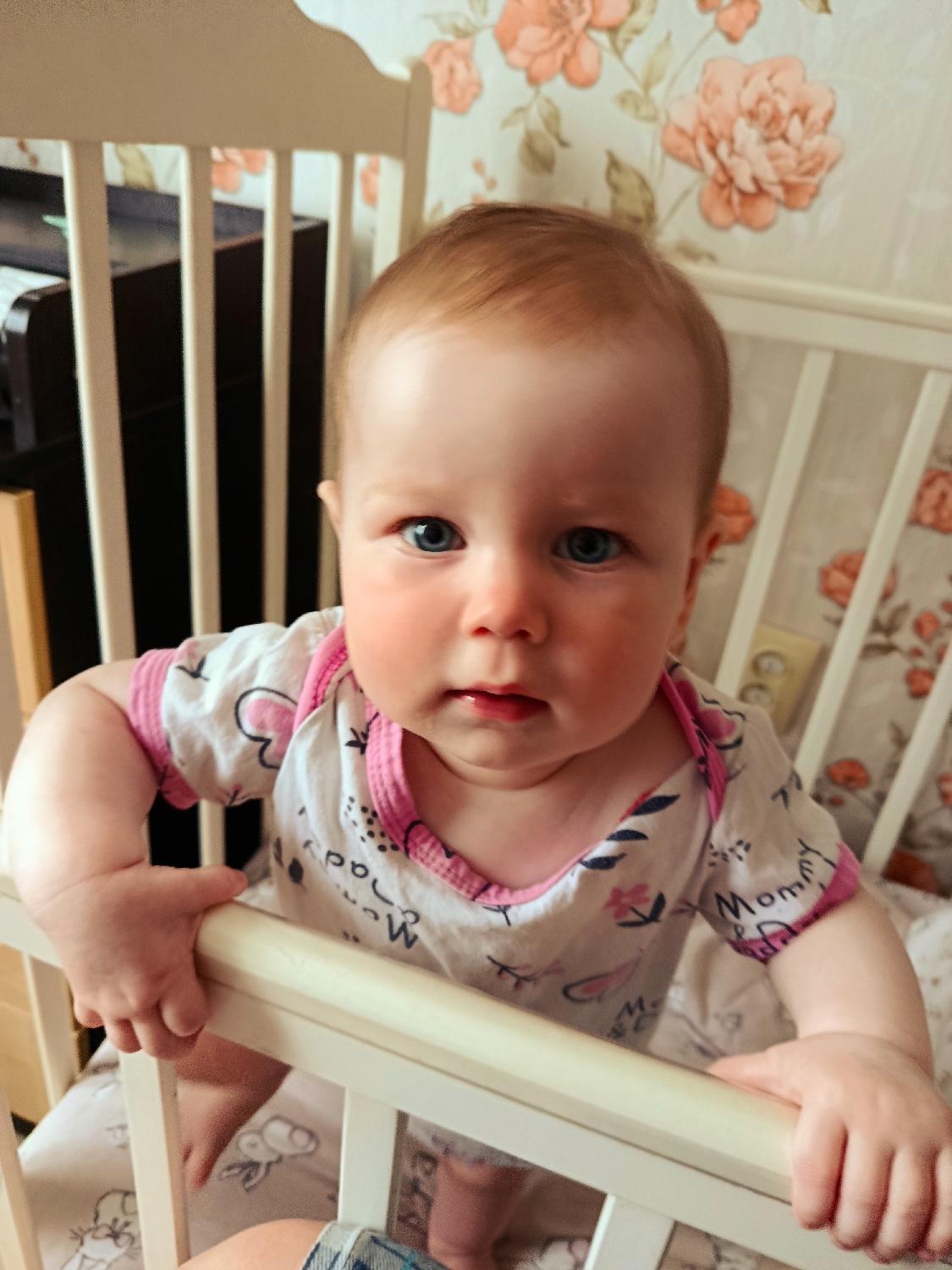 A close-up, eye-level portrait features a baby standing upright inside a white wooden crib, looking directly at the camera. The baby has fair skin, rosy cheeks, bright blue eyes, and light reddish-brown hair. Dressed in a white romper with pink trim, the garment is patterned with hearts, flowers, and the word "Mommy" clearly visible on the right sleeve area.
The baby's hands are gripping the top rail of the crib, indicating a stable standing position. Behind the crib, the wall is covered with wallpaper featuring a prominent floral design of large orange and pink flowers with green leaves. Through the crib slats on the left, a dark piece of furniture is partially visible. On the wall to the right, a light-colored electrical outlet can be seen. Inside the crib, a white sheet or blanket patterned with cartoon rabbit figures is visible. A small portion of a person's leg, clad in blue jeans, is visible at the very bottom of the frame, suggesting an adult is nearby, likely the one taking the photograph. The baby's expression is neutral, with wide, attentive eyes, conveying a sense of curiosity or slight seriousness. The setting appears to be indoors, likely a nursery or child's room, during daytime.