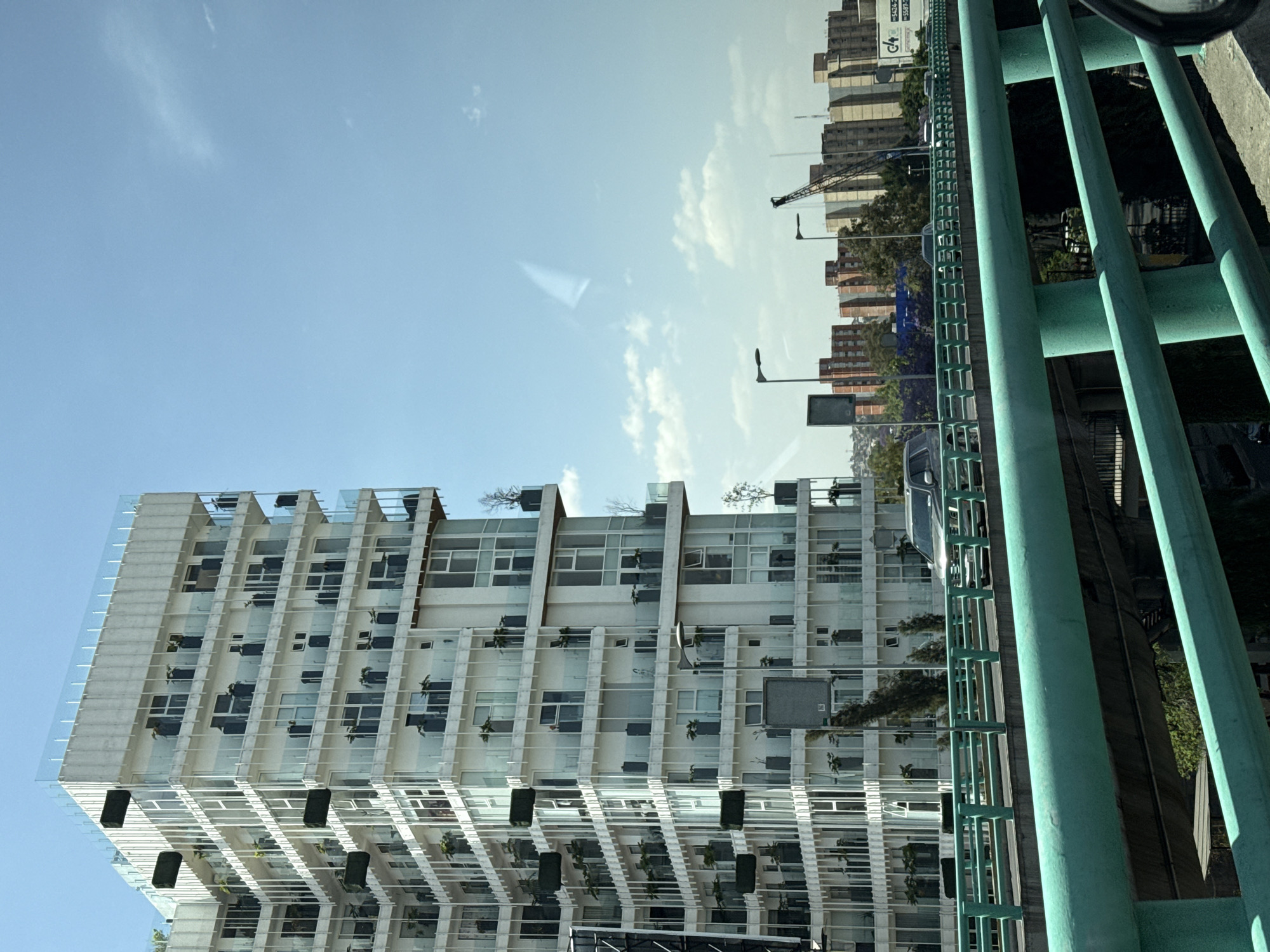 The image is taken from what appears to be a vehicle traveling along an elevated road structure in Mexico City, Mexico.
A tall, white modern building with many windows and balconies is on the left, contrasted against a blue sky with scattered clouds. The balconies appear to have plants or planters.
On the right, green metal guard rails and structural supports partially obscure the view of other buildings further in the background. A crane is visible amongst those buildings.
The photo is taken from an upward angle, looking toward the sky.