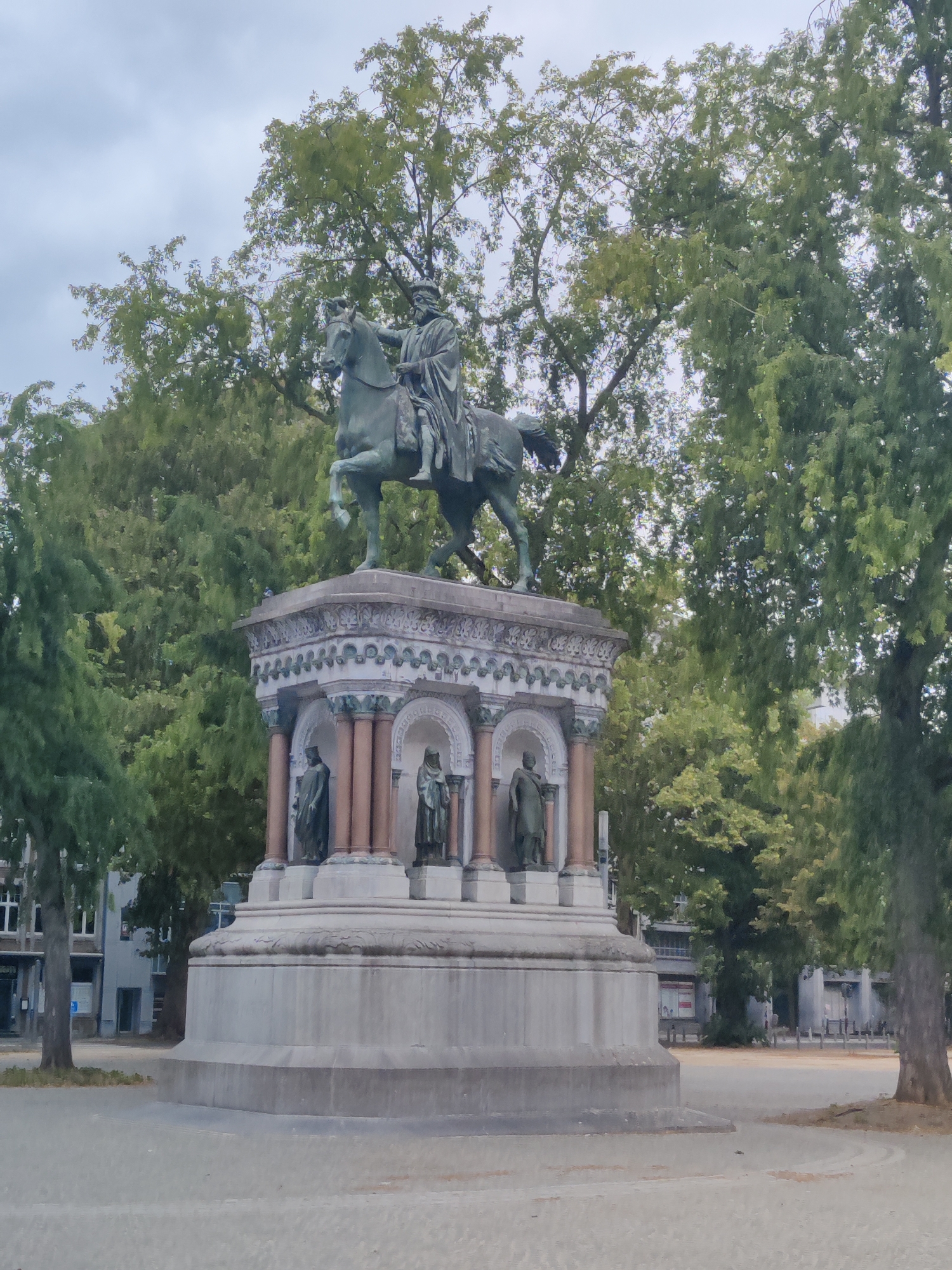 A bronze equestrian statue of King Leopold II stands atop a multi-tiered monument in Place du Théâtre, Liège, Belgium. The king, depicted wearing a cape and crown, sits astride a horse with its front legs raised. The monument's base features decorative elements and alcoves containing smaller statues of robed figures. The scene is framed by lush green trees, with buildings and an overcast sky visible in the background. The ground in front of the monument is paved.