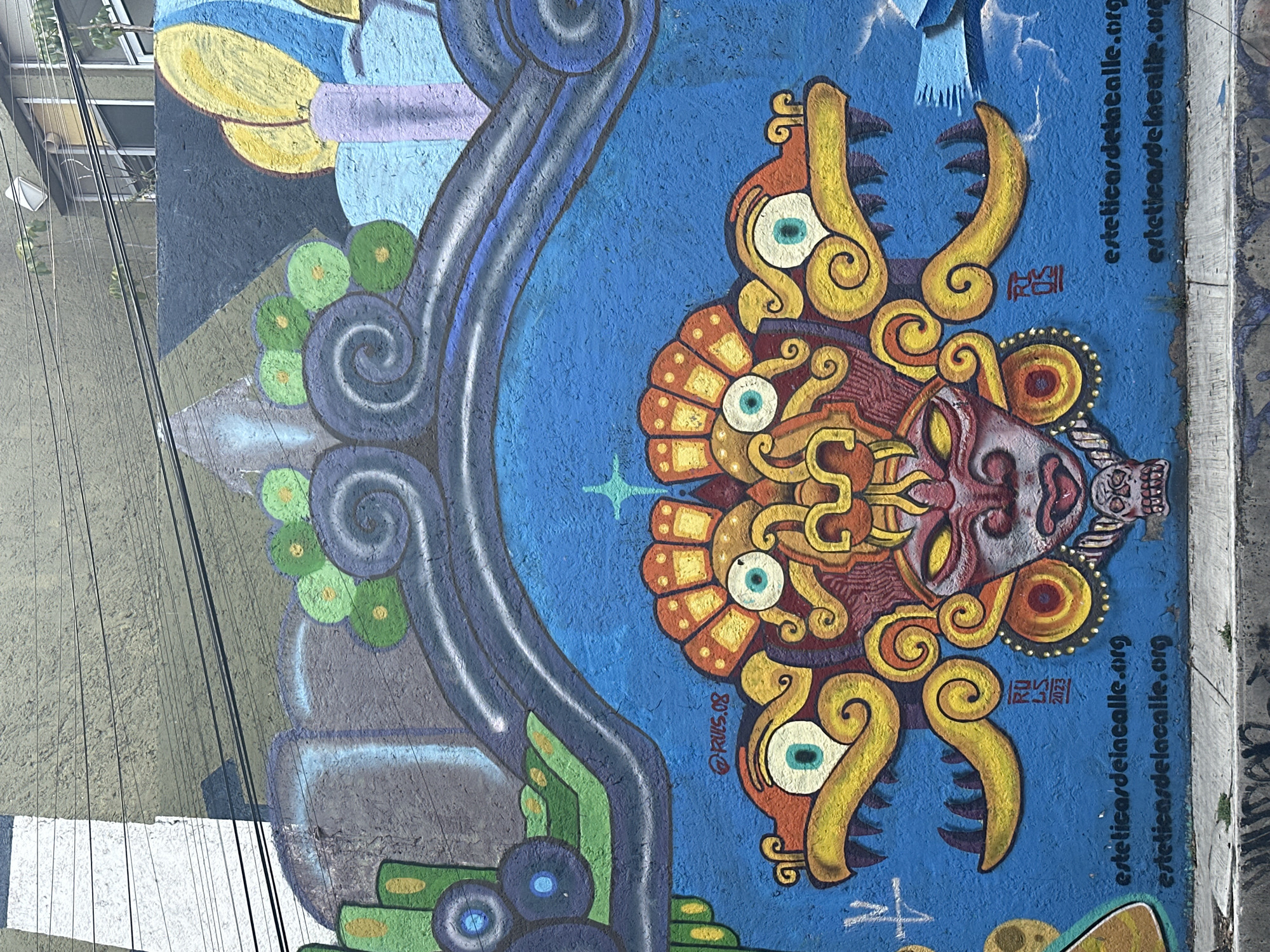The image shows a vibrant mural in Mexico City, Mexico. The mural features a complex, stylized Aztec-inspired mask in the center. The mask is painted in shades of orange, yellow, red, and maroon. It has large, ornate decorations around its eyes and mouth, with swirling, horn-like protrusions. The background is a bright blue color. To the left of the mask, there is a swirling gray and green design. At the top left of the image, glimpses of buildings and utility lines are visible. Text is painted on the right side of the mural, reading "esteticadelacalle.org". A signature on the mural appears as "RWS.08" and "RWS 2023" indicating it was created in 2008 and updated in 2023. The photograph is taken from a slightly elevated angle, looking down at the mural. The condition of the mural appears to be generally good.