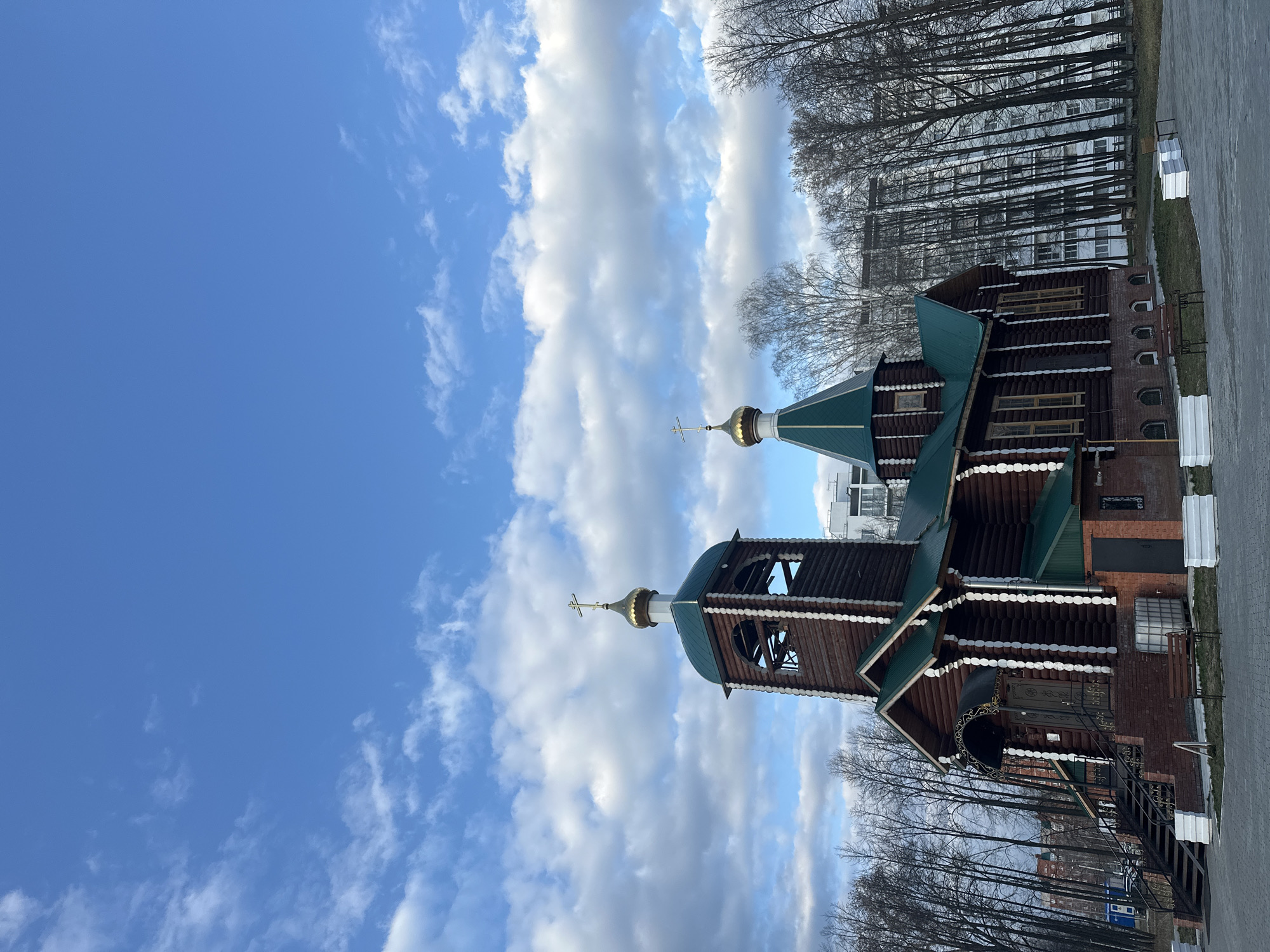 The image, taken in Chaikovsky, Russia, showcases a traditional wooden building, likely an Orthodox church or chapel, set against a vibrant blue sky dotted with white, fluffy clouds. The structure is characterized by its dark brown log walls, accented with decorative white trim, and features prominent green roofs.
Two distinct cupolas, both topped with golden onion domes and Orthodox crosses, are visible. The taller structure on the left appears to be a bell tower with open arches, while a smaller dome is situated further to the right. Bare deciduous trees, suggesting late winter or early spring, form a dense backdrop, partially obscuring a large, light-colored multi-story building in the distance.
In the foreground, a paved path or road runs along the right side, accompanied by several white rectangular objects, possibly benches or planters. Details from the provided crops show intricate sections of the building, including a brick wall with arched recesses and a dark entrance door adorned with ornate metalwork, accessible via a set of stairs. The scene is brightly lit, indicating daytime, and conveys a serene and peaceful atmosphere. No specific activity or legible text is visible.